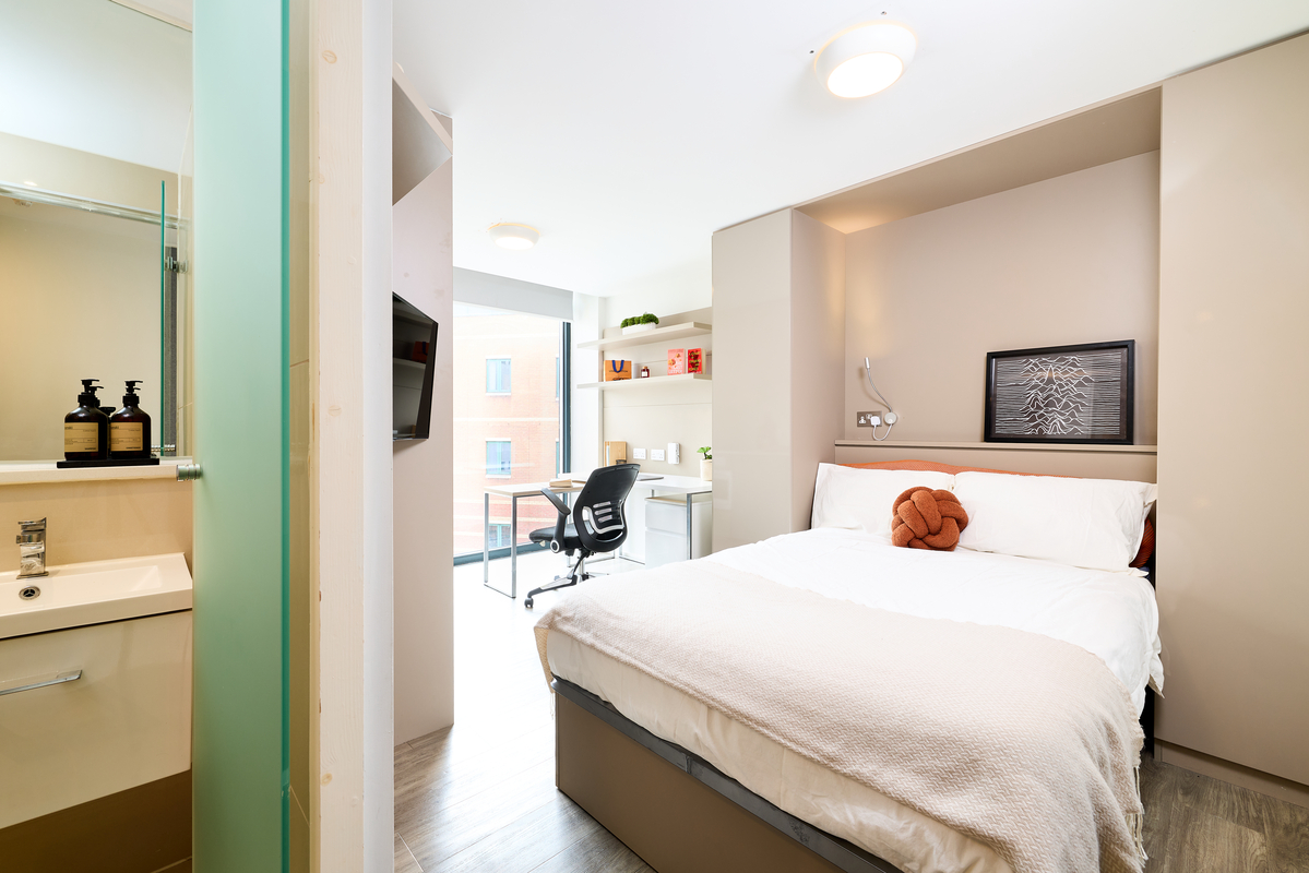 A modern, well-lit Portland House bedroom with a double bed, white bedding, a small desk by the window, shelves with books, a wall-mounted TV, and an adjoining bathroom with a sink and toiletries.