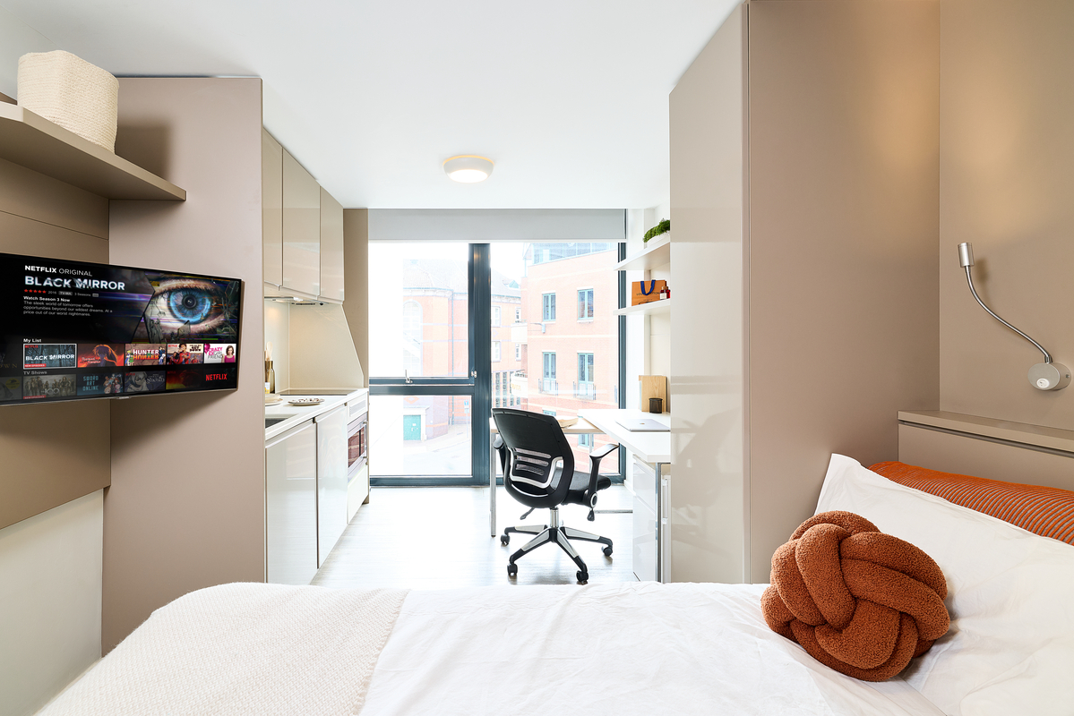 A modern studio apartment in Portland House with a bed, orange knotted pillow, wall-mounted lamp, desk and chair by a window, kitchenette, and a TV showing Black Mirror on Netflix. Bright, minimalist decor with ample natural light.
