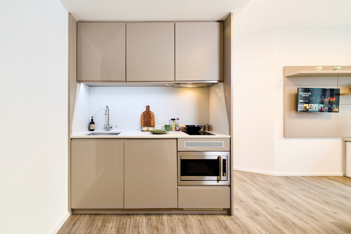 Modern, compact kitchen with beige cabinets, a sink, a built-in microwave, a cooktop, and a wooden countertop with kitchen items. Light wood flooring and a wall-mounted TV are visible in the background.