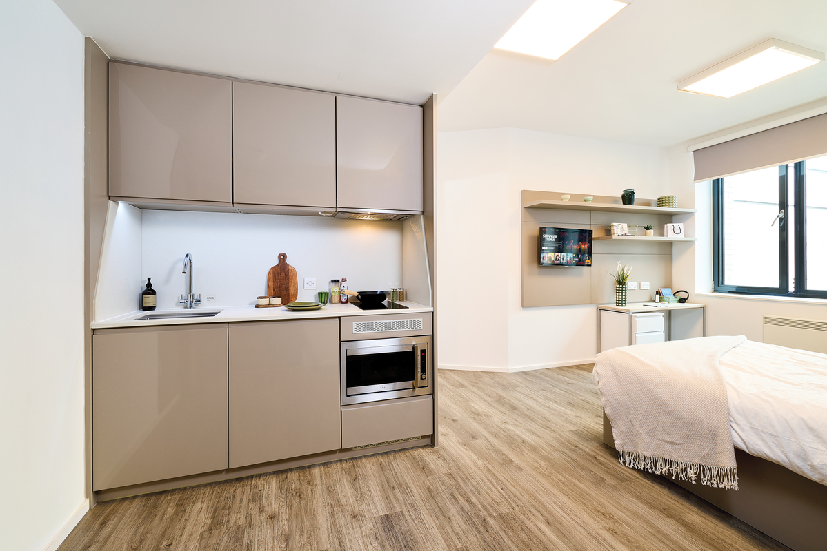 Modern studio apartment with a sleek gray kitchenette, sink, microwave, and storage; bed with white bedding nearby; wall shelves and TV opposite a large window letting in natural light.