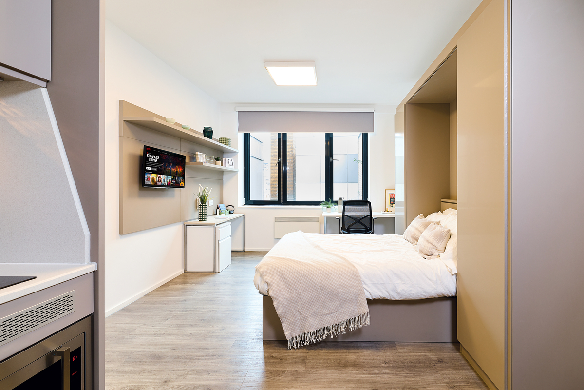Modern studio apartment with a neatly made bed, desk by the window, wall-mounted TV, shelves with decor, and light wood flooring. Room is bright with natural light from large windows.
