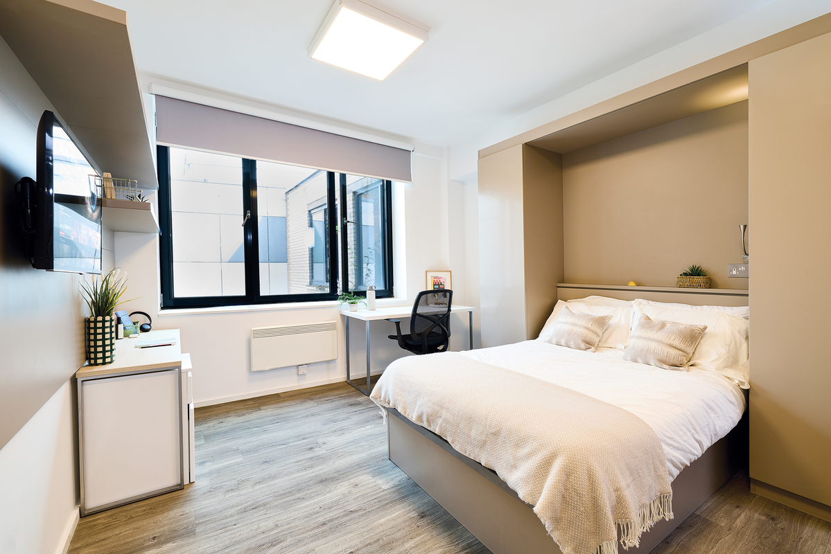Modern, tidy studio apartment with a double bed, white bedding, built-in shelves, a wall-mounted TV, a small fridge, a desk and chair by the window, and light wood floors. Natural light fills the room.