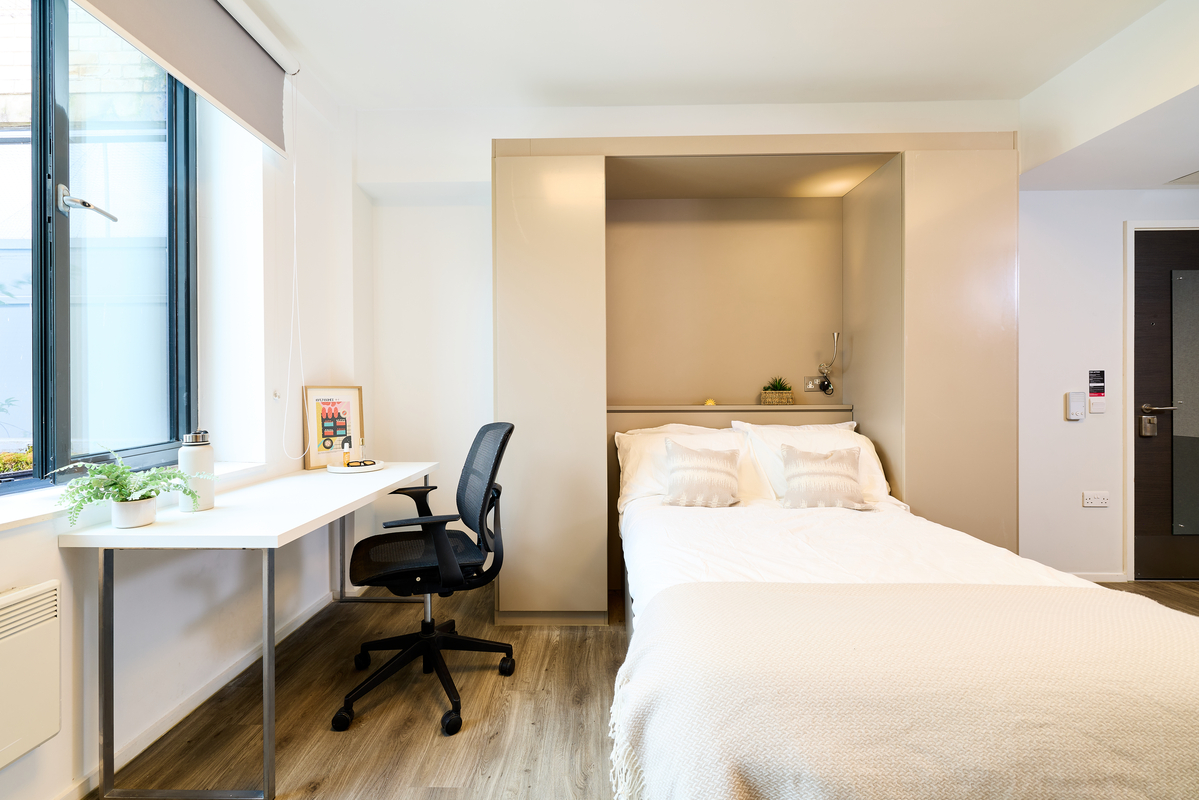 A modern, bright bedroom features a double bed with white bedding, a built-in headboard, a small desk with a chair by a large window, and wood flooring. Decor includes plants and minimal accessories.