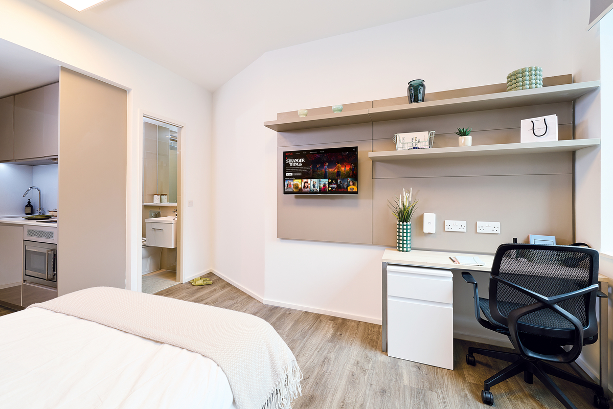 A modern, tidy bedroom with a bed in the foreground, a wall-mounted TV, shelves with decor items, a white desk and office chair, and an en-suite bathroom visible through an open door.