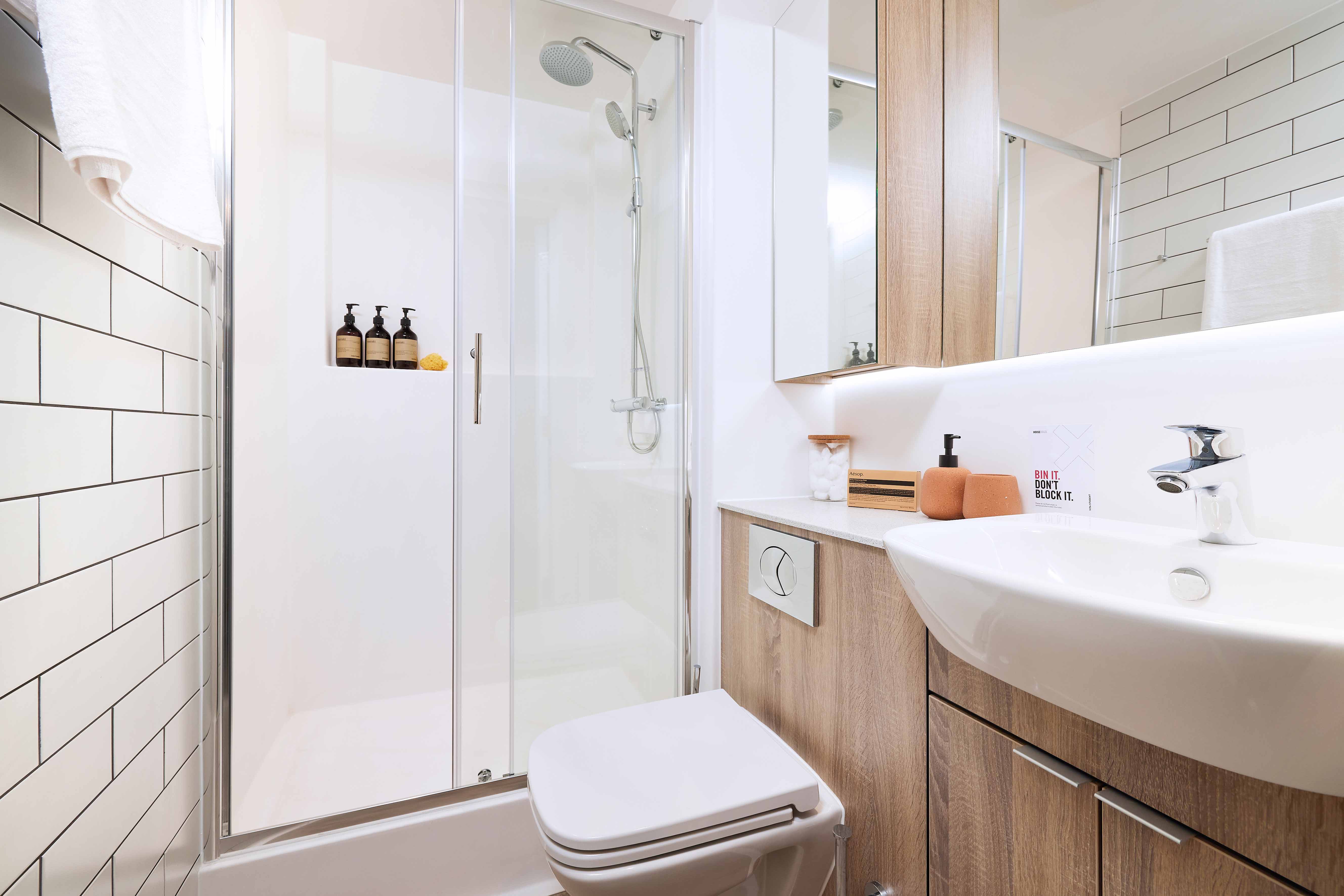 A modern bathroom with a glass-enclosed shower, white subway tiles, wooden cabinets, and a white sink. There are soap dispensers in the shower and a towel on a bar. The space is bright and neatly organized.