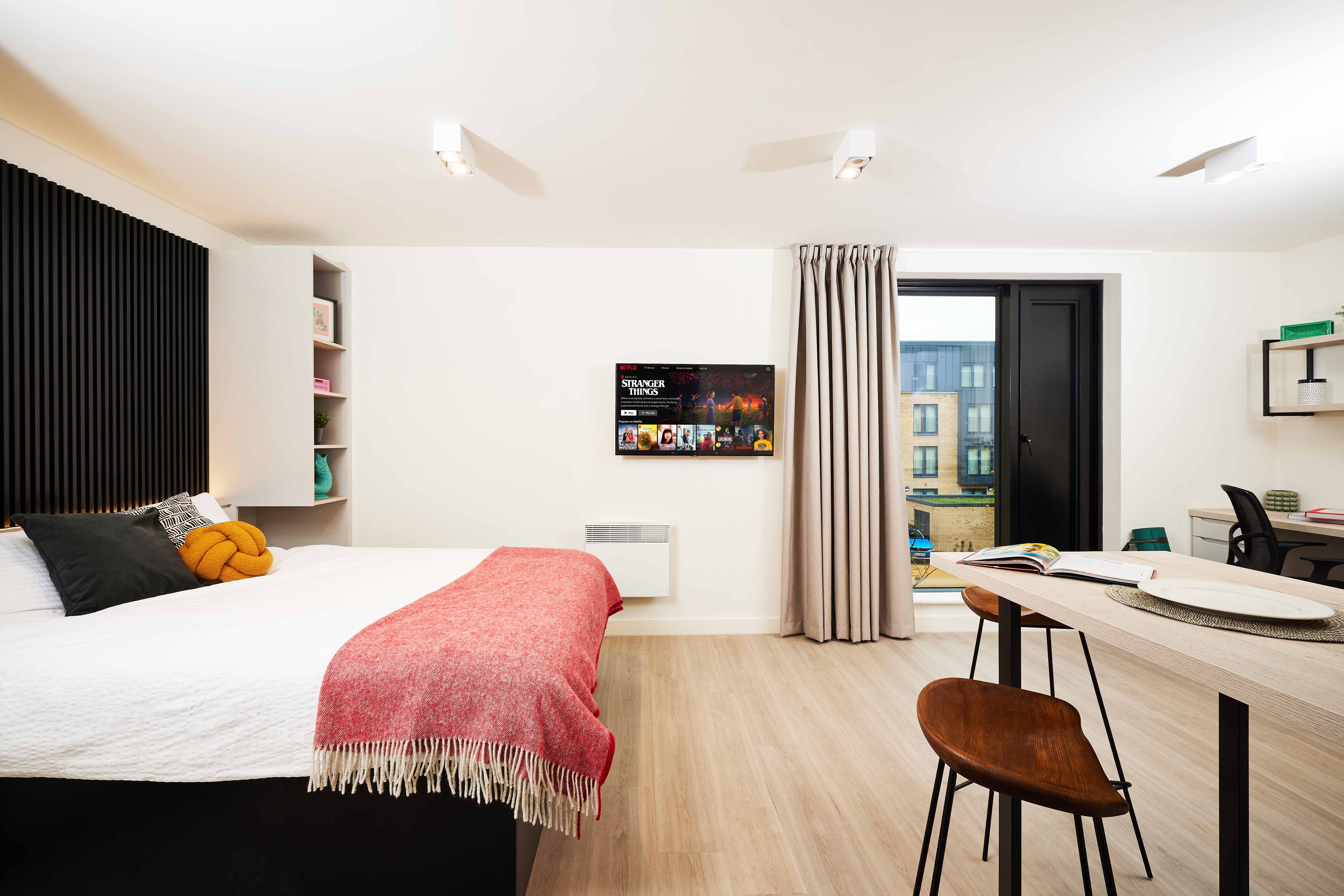A modern studio apartment with a neatly made bed, red throw blanket, and a small orange pillow. There’s a TV on the wall, a desk and chair by the window, and a balcony door with a curtain. Light wood flooring and minimal decor create a cozy ambiance.