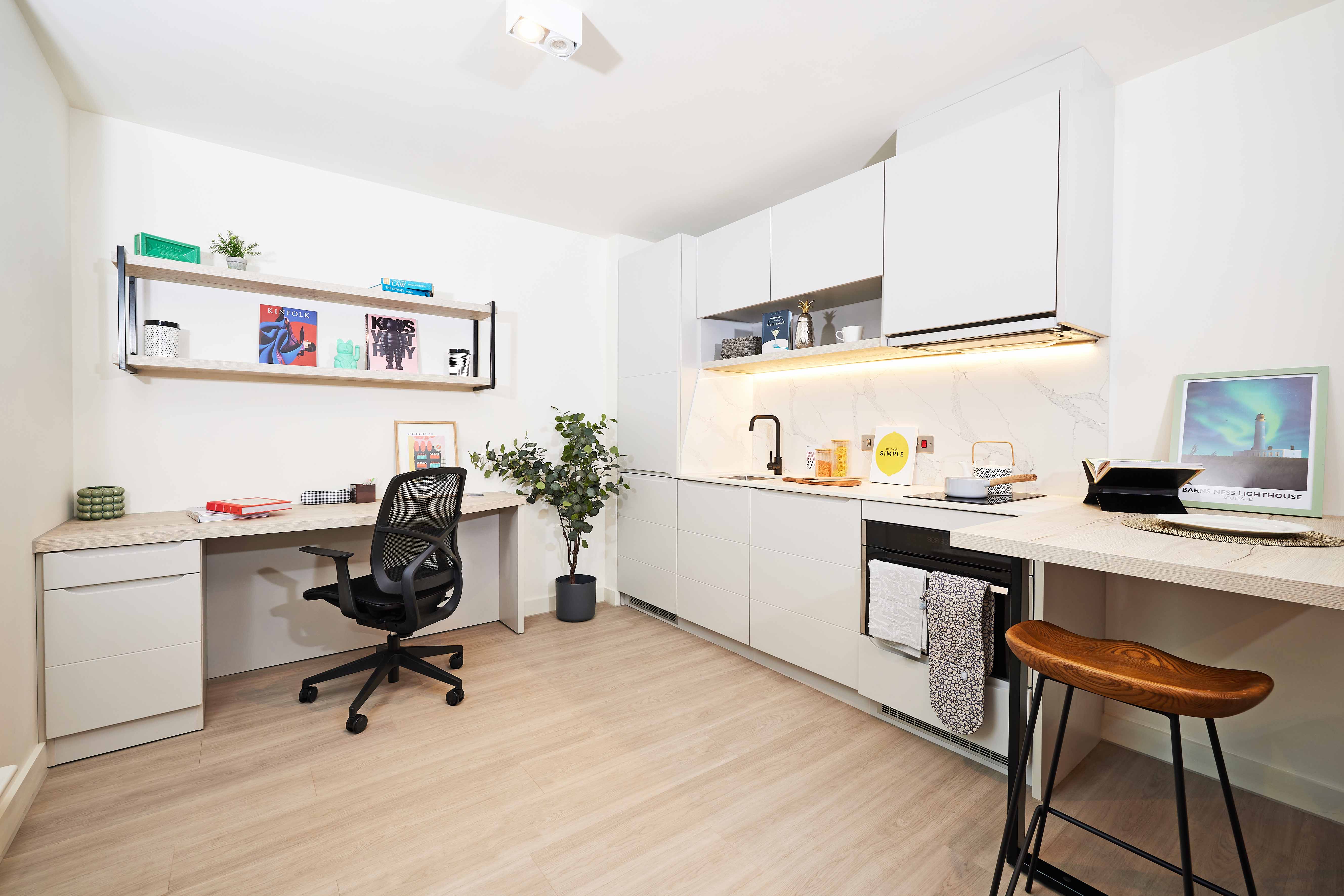 Modern studio apartment with a kitchenette and workspace. Features include white cabinets, a built-in desk with a black chair, wall shelving with books and decor, a plant, and a bar stool at a wooden counter. Light wood flooring throughout.