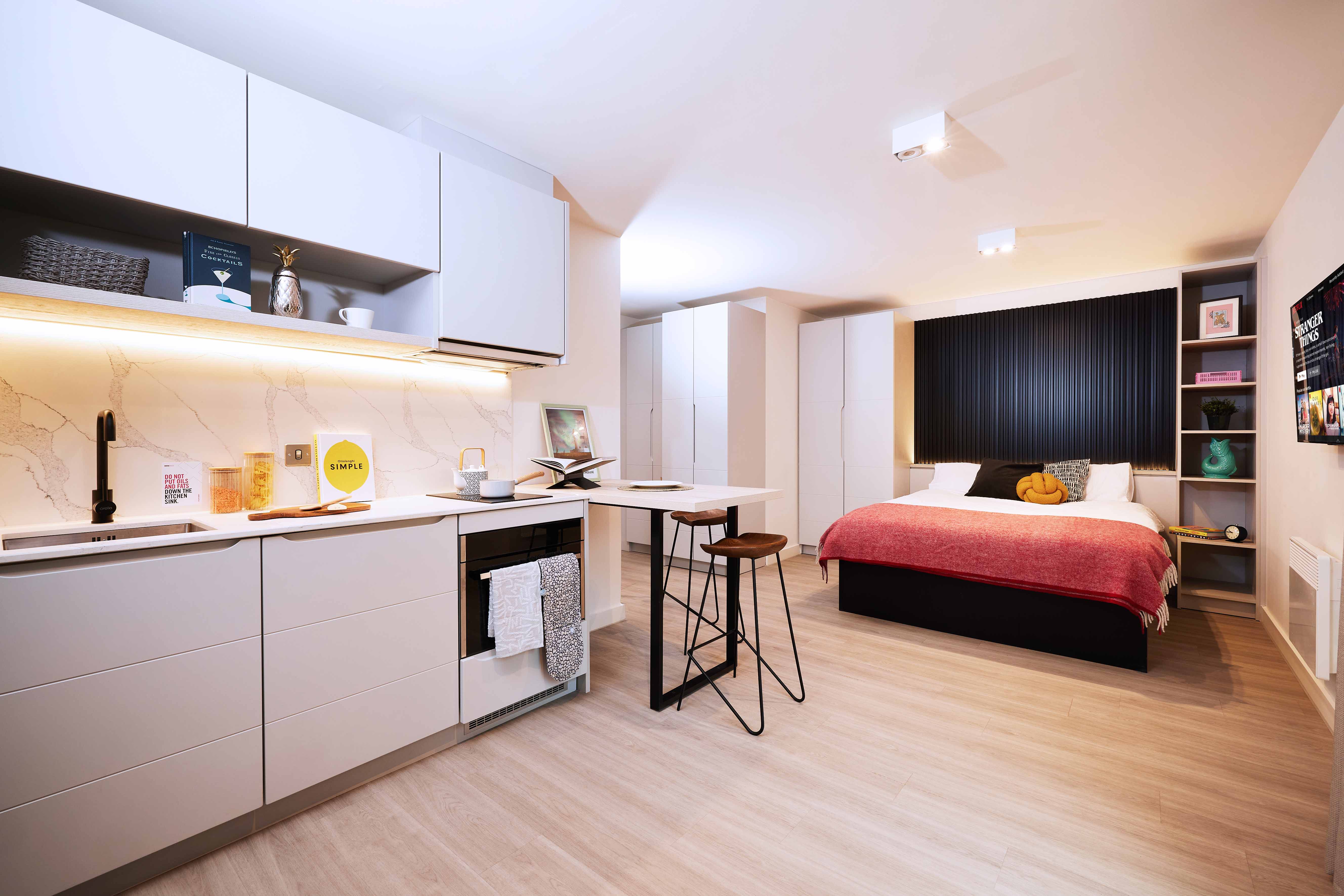 Modern studio apartment with a kitchenette and a bed. The kitchen has white cabinets, a countertop with a cooktop, and a bar stool. The sleeping area features a double bed with a red blanket, pillows, and a decorative black panel behind. Bright lighting.