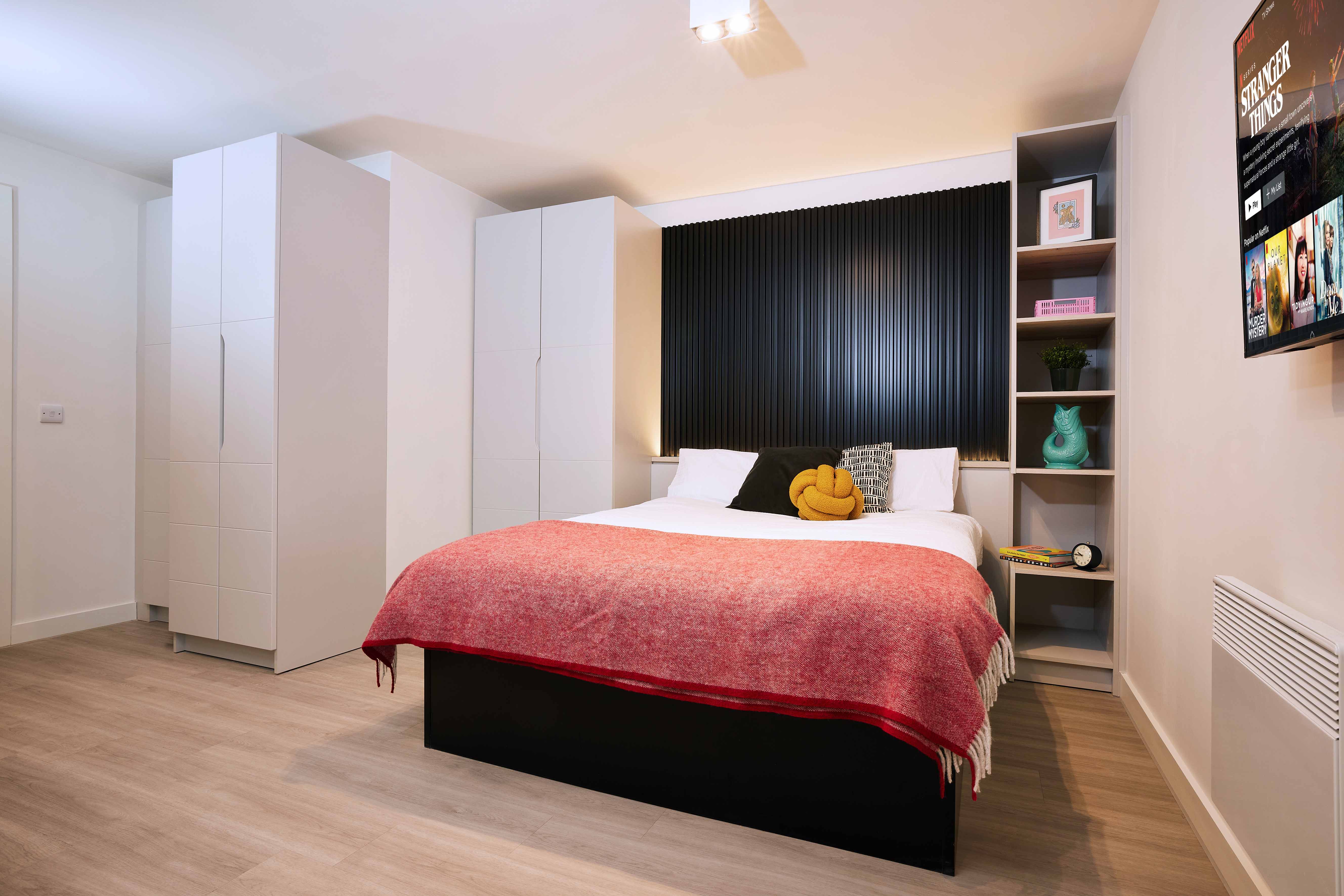 Bedroom with a double bed featuring red and white bedding. A modern black headboard is flanked by white wardrobes and shelves. Decor includes books and colorful accents. A wall-mounted TV is on the right. Light wood flooring enhances the contemporary look.