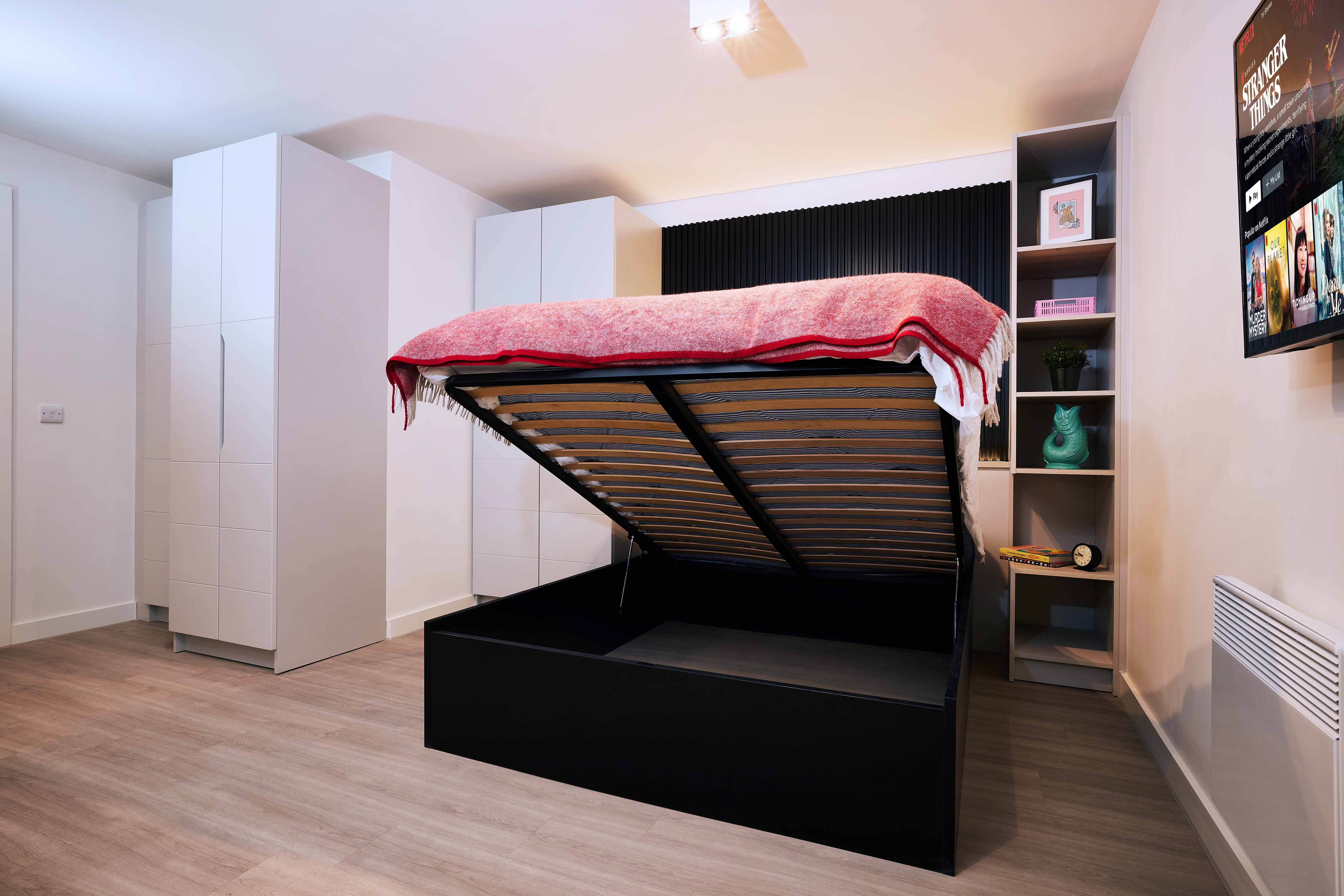 Modern bedroom with a raised ottoman bed revealing storage space underneath. The bed is topped with a pink blanket. White wardrobes, a small shelf with decorations, and a wall-mounted TV are visible against neutral-toned walls and flooring.