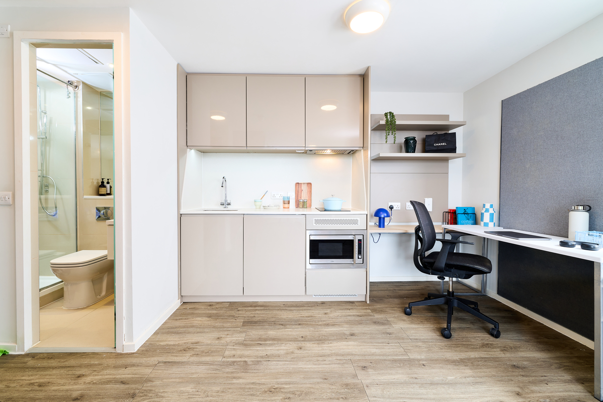 Modern studio apartment at Portland House featuring a small kitchen with microwave and sink, a work desk with chair, shelves, wood-style flooring throughout, plus a view into the bathroom with shower and toilet.
