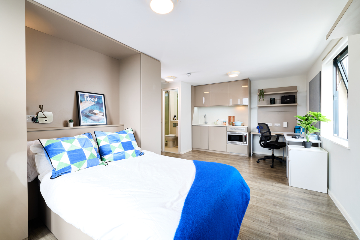 A modern studio apartment at Portland House with a double bed, geometric-patterned pillows and blue blanket, kitchenette, ensuite bathroom, desk and chair, potted plant, and a large window filling the space with natural light.
