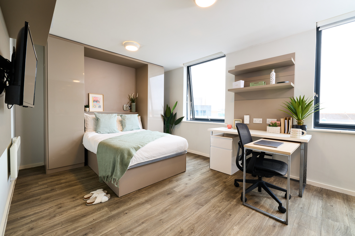 A modern, bright bedroom at this Portland accommodation features a neatly made bed with green accents, a desk and chair by the window, shelves with decor and books, houseplants, and light wood flooring filled with natural light.