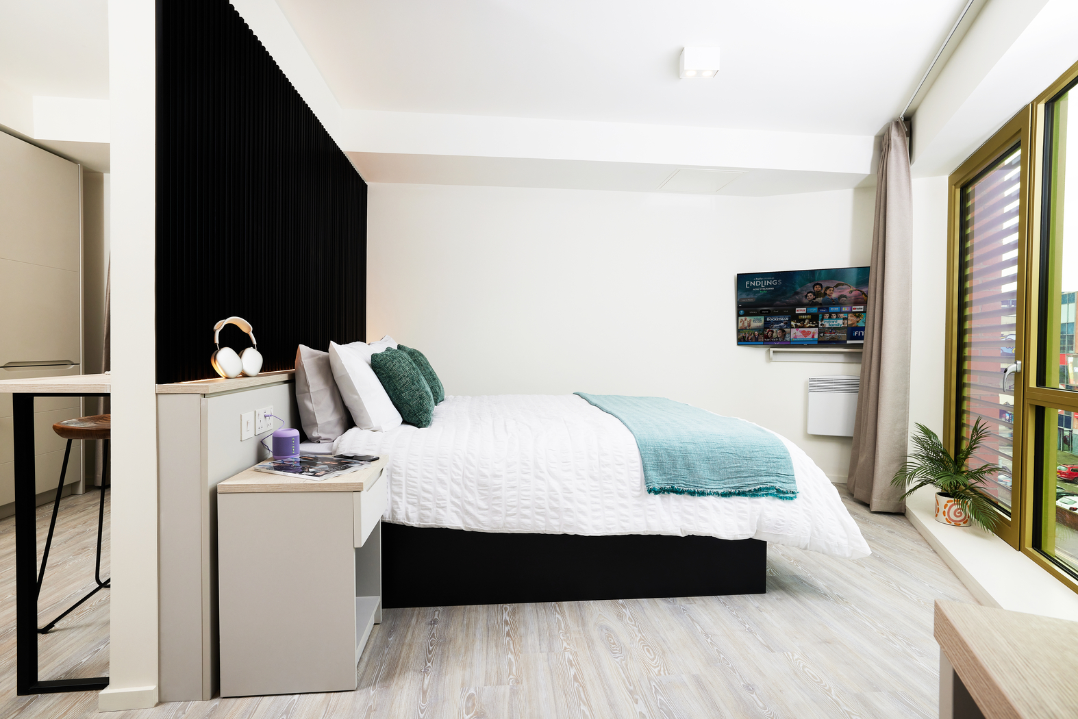 A modern bedroom on Bruce Street showcases a neatly made bed with white and teal bedding. A small nightstand holds a lamp and books. The space features a black accent wall, floor-to-ceiling windows with curtains, light wood flooring, and a wall-mounted TV for added comfort.