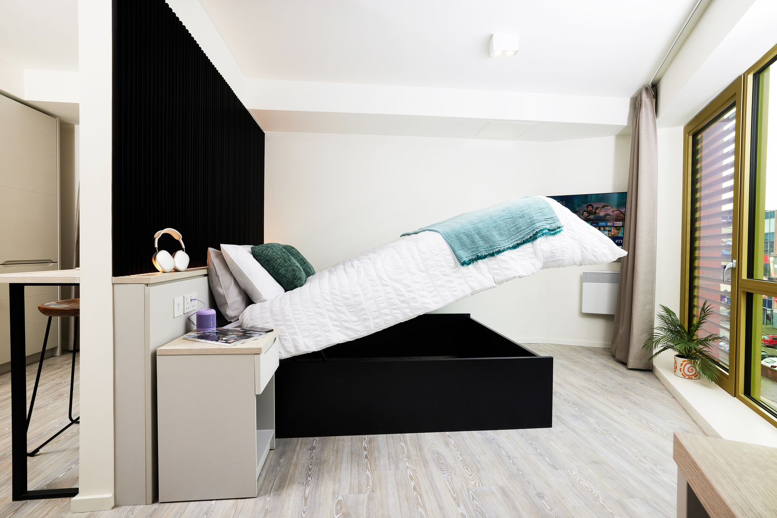 A modern bedroom on Bruce Street features a bed with an elevating mattress mechanism, revealing storage space underneath. The room showcases a minimalistic design with a black accent wall, wooden flooring, a bedside table, and a large window offering an inviting view outside.