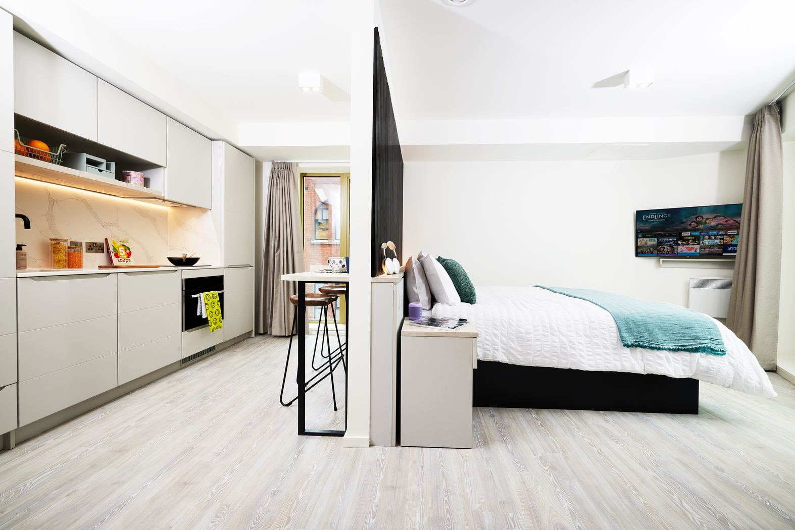 A modern studio apartment on Bruce Street offers a partitioned layout, separating the kitchen and bedroom. The kitchen boasts light cabinetry, a stovetop, and countertop. The bedroom features teal accents with a made bed and wall-mounted TV, all set over elegant light wood flooring.