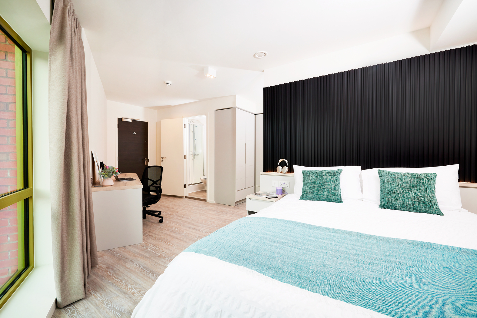 The modern bedroom on Bruce Street features a large bed with teal accents on the bedding. A desk and chair are set against a wall, next to a wardrobe and an open door to the bathroom. Natural light filters in through the window adorned with a curtain, completing this serene space.