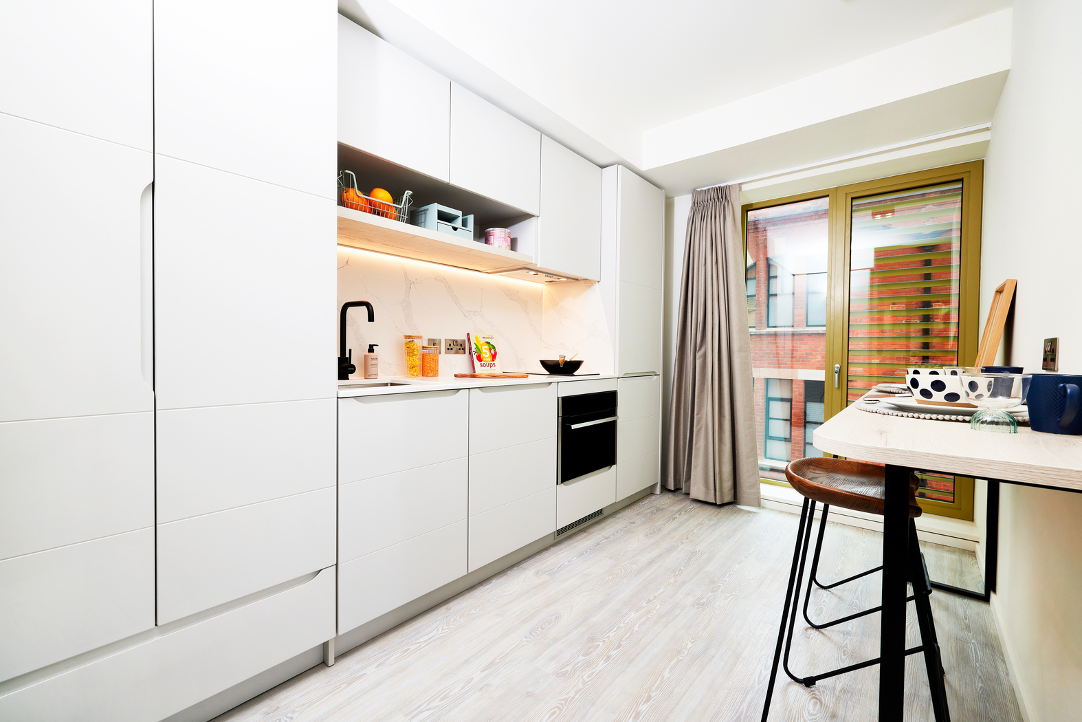 Located on Bruce Street, this modern kitchen features white cabinetry, wood countertops, and black accents. It includes a built-in oven, open shelving with dishware, and a sink. A small breakfast bar with stools sits on the right, while large windows with curtains grace the background.