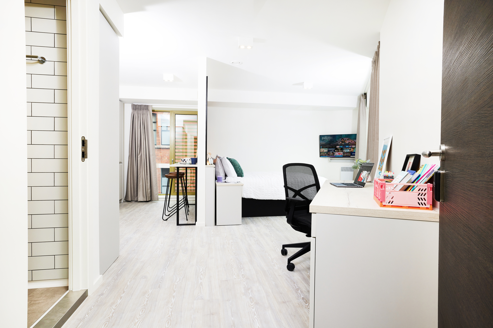 A modern, well-lit studio apartment on Bruce Street boasts white walls and light wood flooring. It features a bed, desk with computer, chair, wall-mounted TV, and a small dining table. A bathroom door is ajar on the left. A large window lets in natural light.