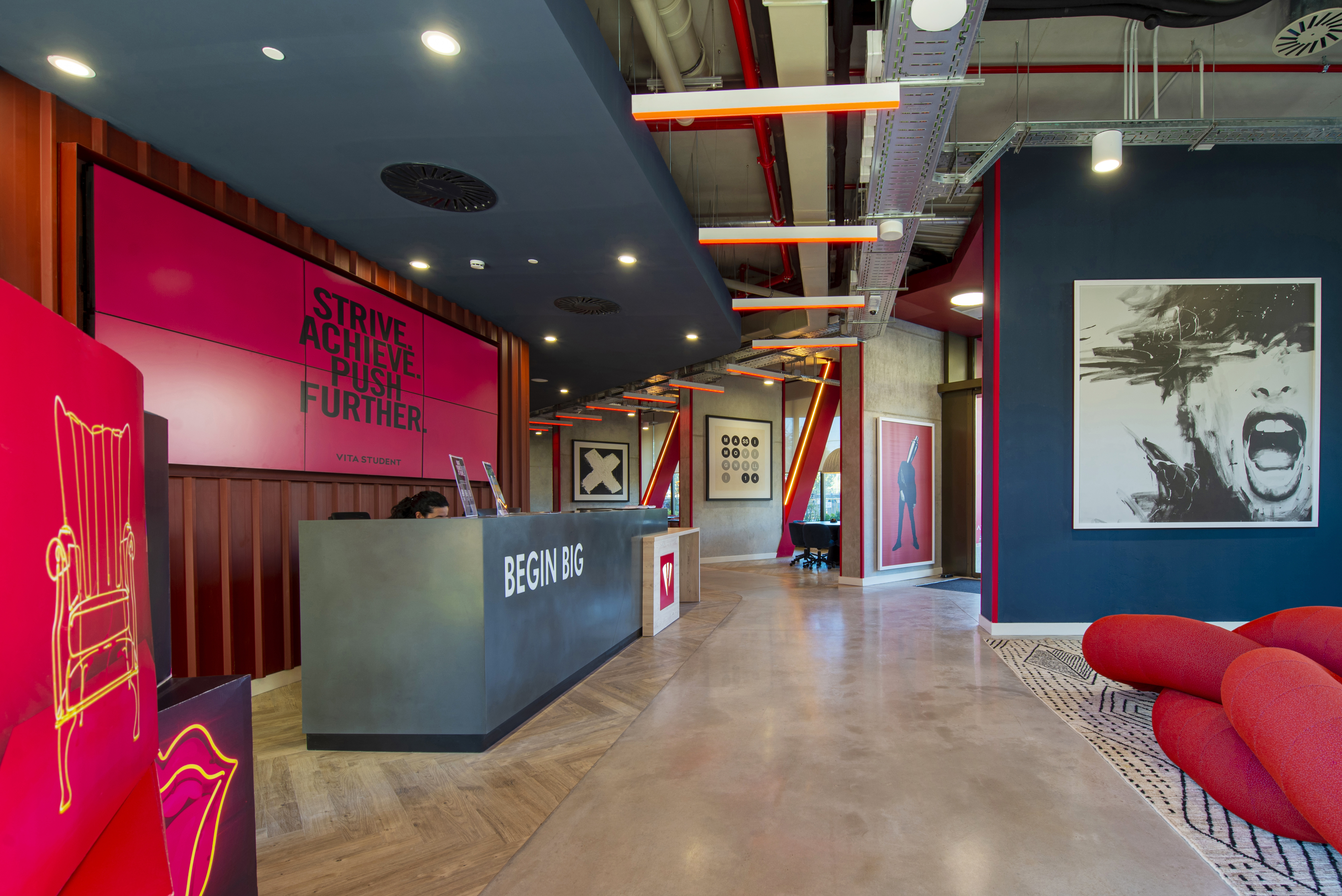 Modern office lobby with a reception desk displaying "BEGIN BIG." Bright red and pink decor, including motivational signs and abstract art. Ceiling with exposed piping and ambient lighting. Red seating area with geometric patterns on the flooring.