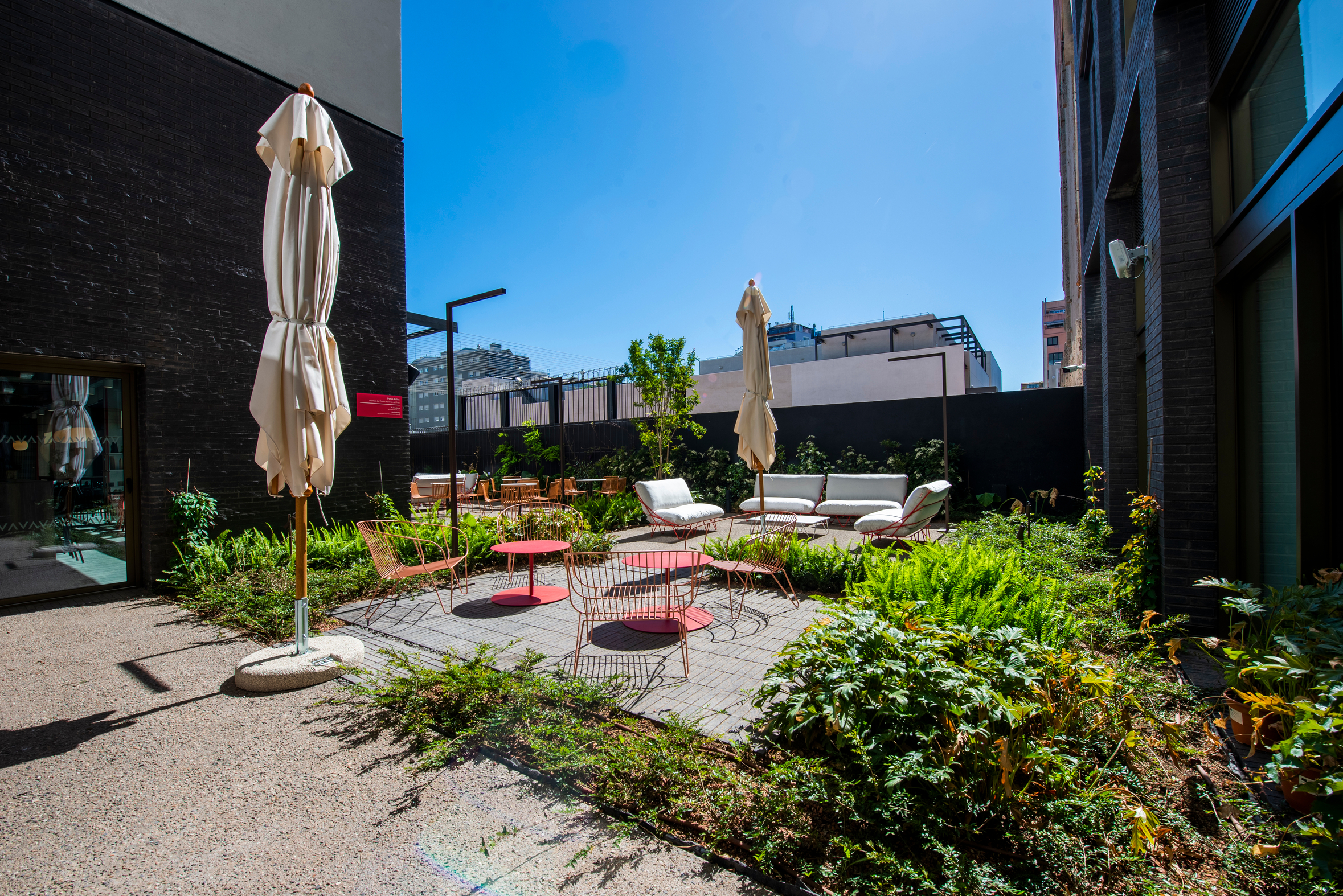 Urban outdoor patio with red tables, white chairs, and umbrellas surrounded by green plants. Brick walls and tall buildings in the background under a clear blue sky.