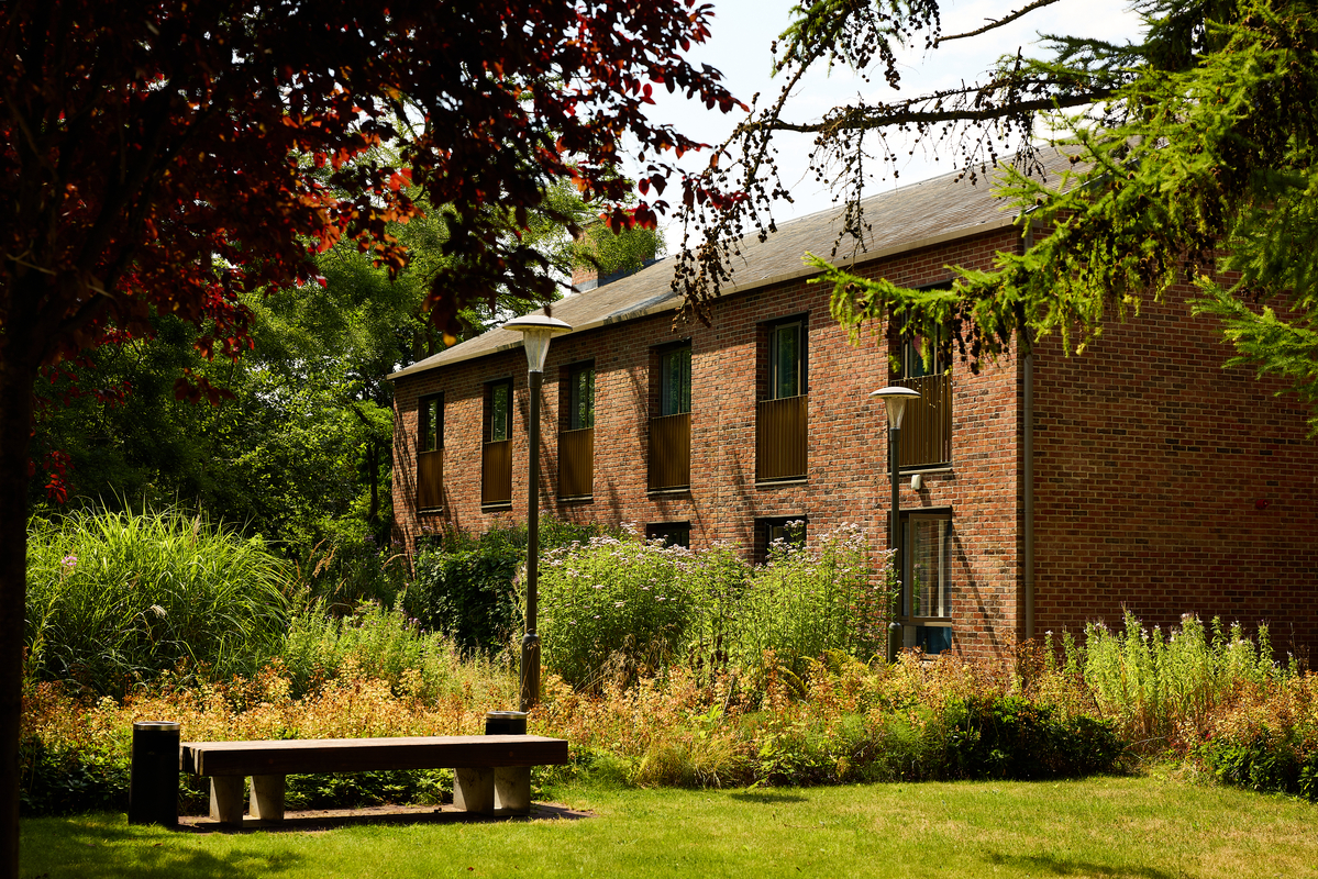 A brick building with two stories is partially shaded by trees. In front, there is a grassy area with a concrete bench surrounded by bushes and plants. The sky is partly cloudy, and the scene has a serene, summery feel.