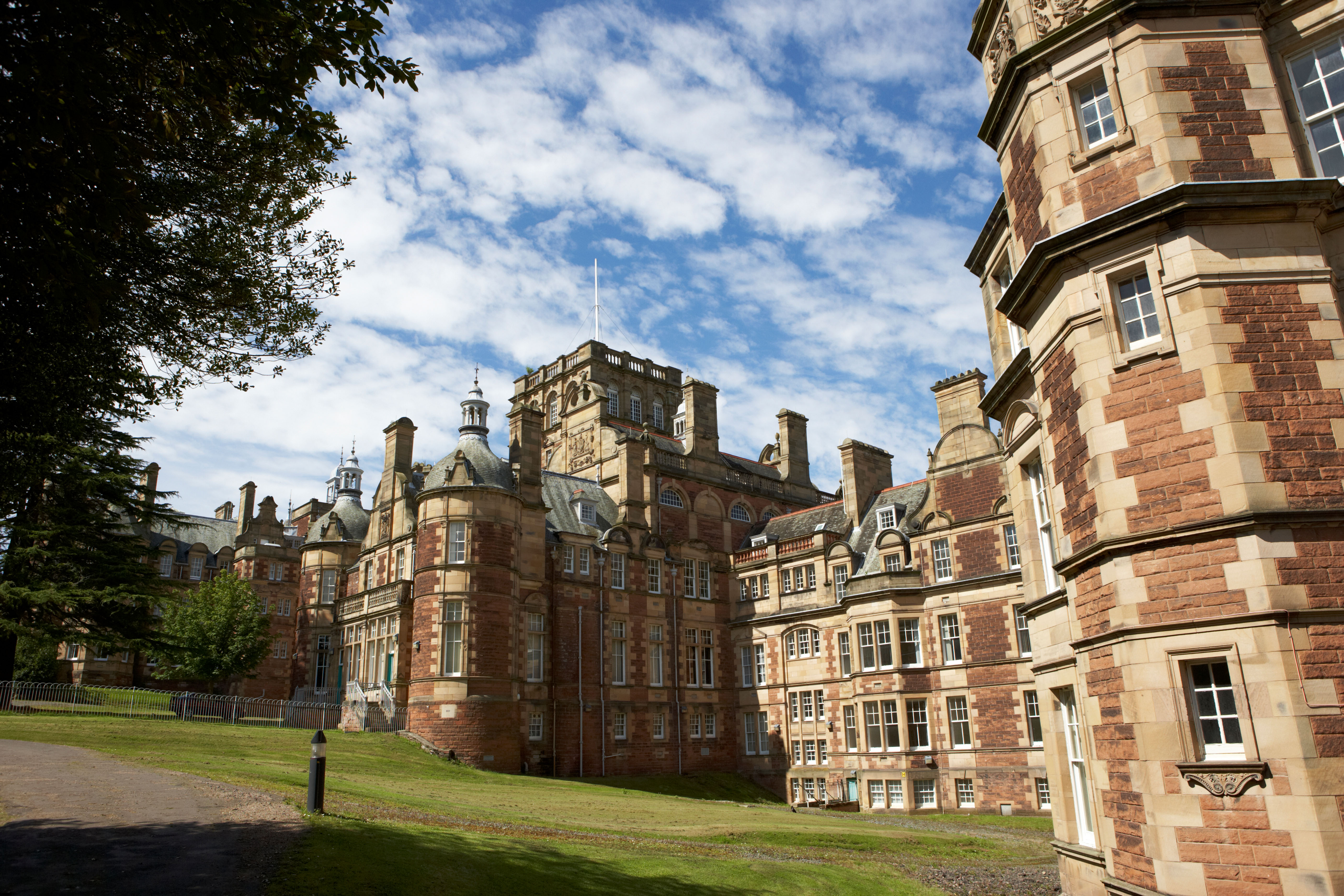 A historic building with red brick and stone exterior, featuring multiple towers and chimneys, under a partly cloudy sky. The foreground has a manicured lawn and a tree on the left side.