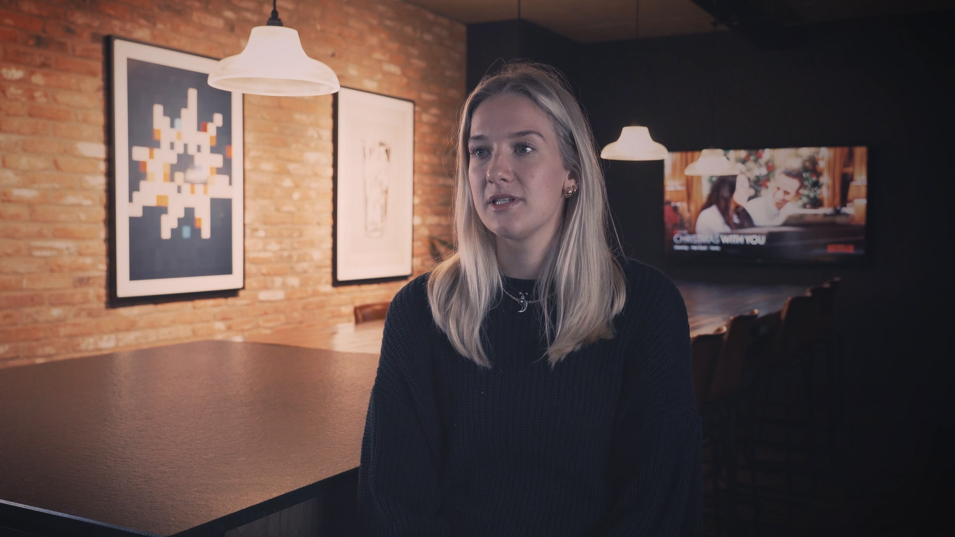 A woman with long blonde hair sits at a table in a cozy student accommodation in Coventry, dimly lit with brick walls. She dons a dark sweater. Framed artworks adorn the wall, and a TV hums softly in the background.
