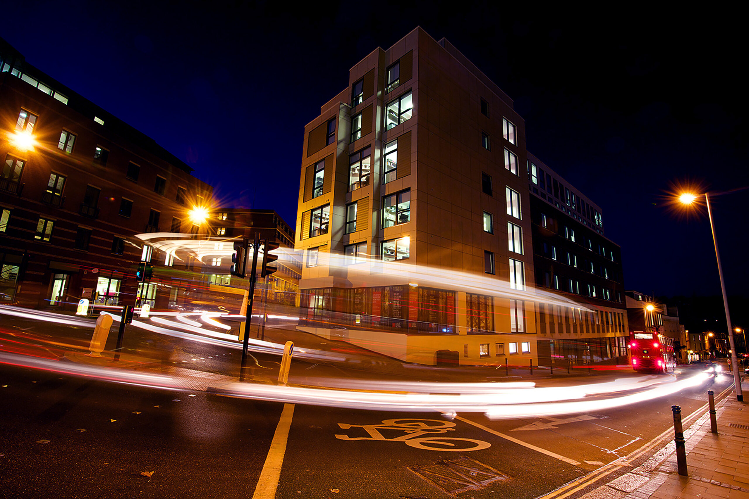 City street at night with light trails from moving vehicles. At the center, Portland House, a modern, illuminated multi-story building, stands tall amidst streetlights and other buildings. In the foreground, a bicycle lane symbol adds dynamic contrast to this urban scene.