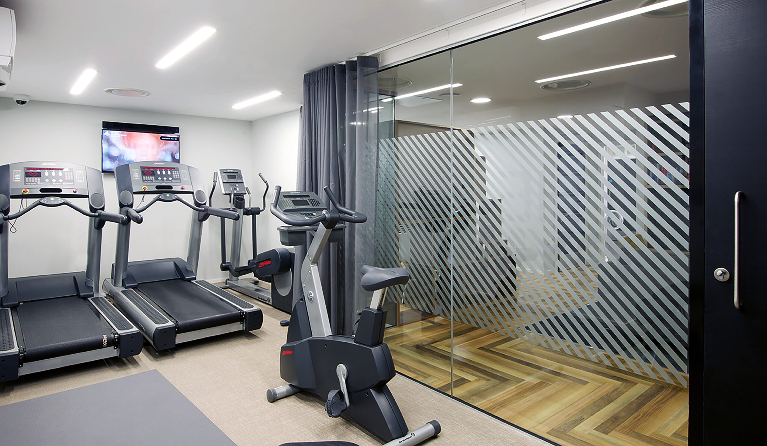 Portland House features a small gym with two treadmills and an exercise bike facing a mirrored wall with diagonal frosted stripes. A wall-mounted TV sits above the treadmills, complemented by sleek overhead lighting. The flooring is a sophisticated blend of laminate and carpet.