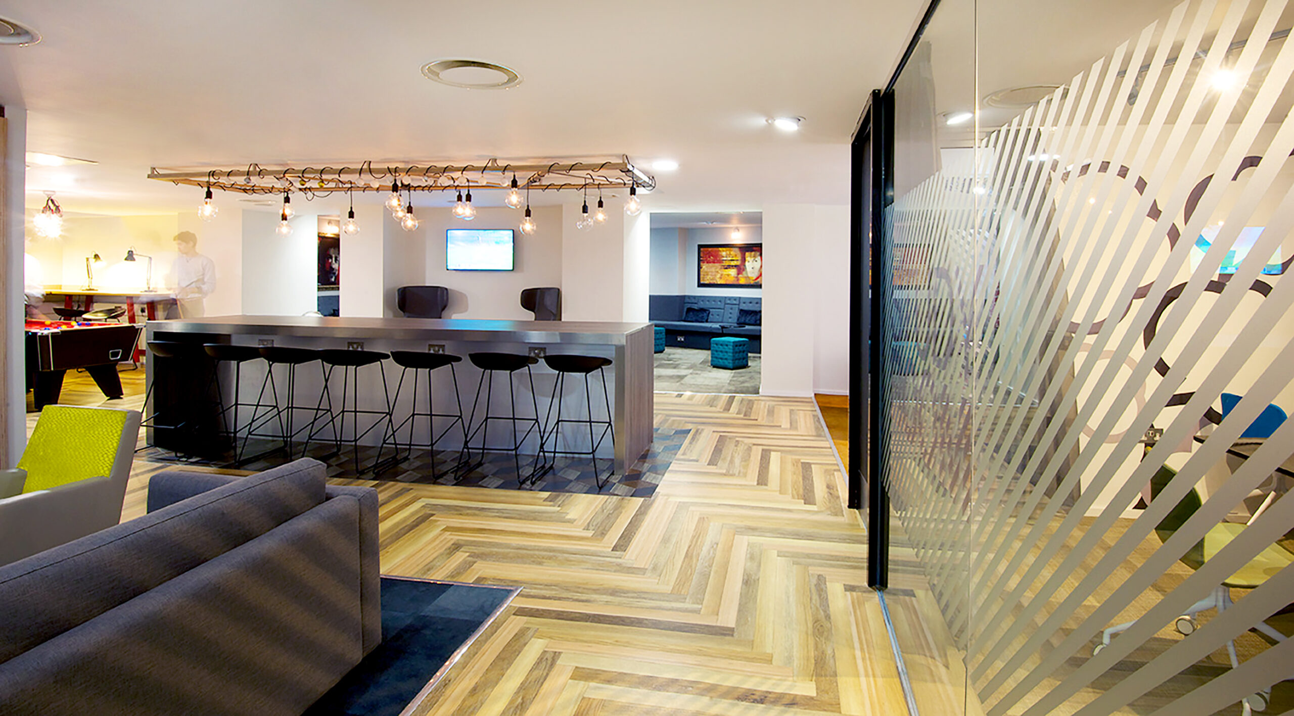 At Portland House, the modern office lounge boasts a central high-top table with barstools on a zigzag wood-patterned floor. Glass walls showcase diagonal stripes, while cozy couches and a TV create a relaxed seating area alongside an inviting game table setup.