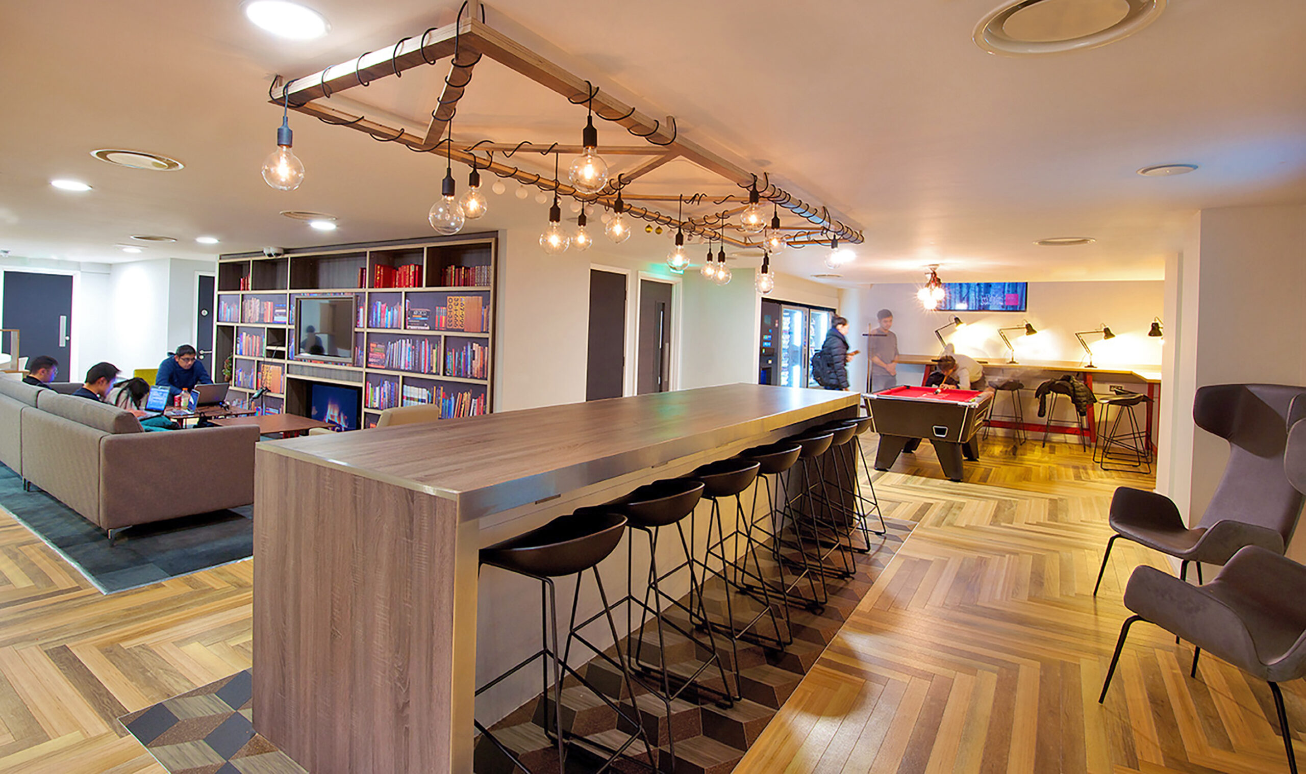 In Portland House, a modern lounge area showcases wooden herringbone flooring and a long high table with black stools. Cozy seating areas, bookshelves, and a pool table fill the background. Warm lighting with exposed bulbs casts an inviting glow overhead.