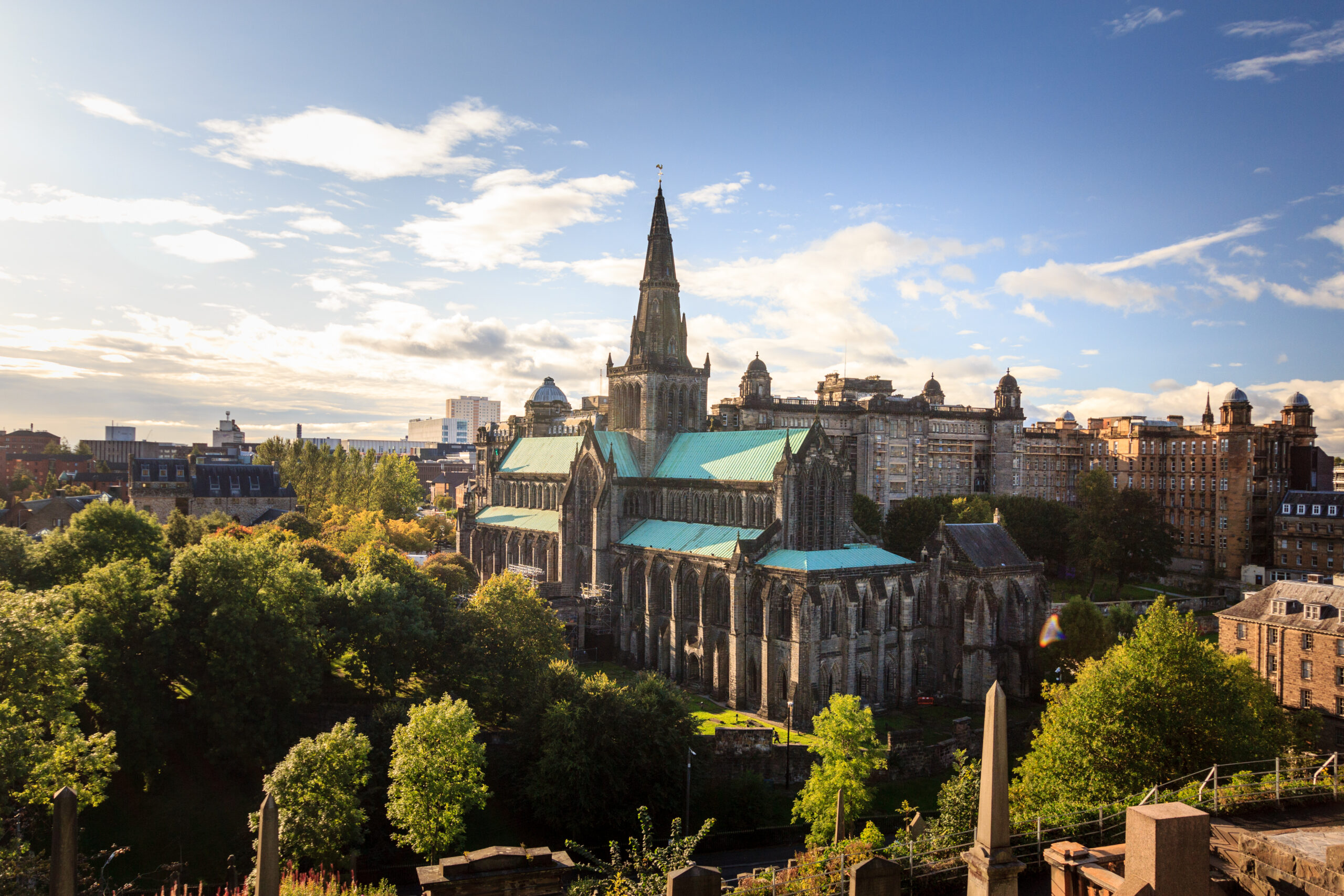 View of a Gothic-style cathedral surrounded by trees and gravestones under a blue sky with clouds. The cityscape is visible in the background, and sunlight illuminates the cathedral's roof and spire.
