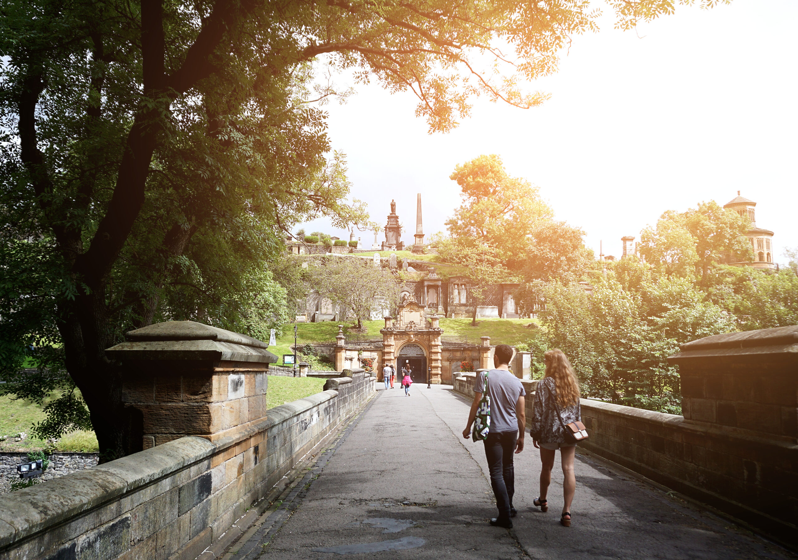 A couple walks down a stone path under arching trees in a sunlit park. They are approaching an ornate entrance with statues and trees in the background. The scene is peaceful and picturesque.