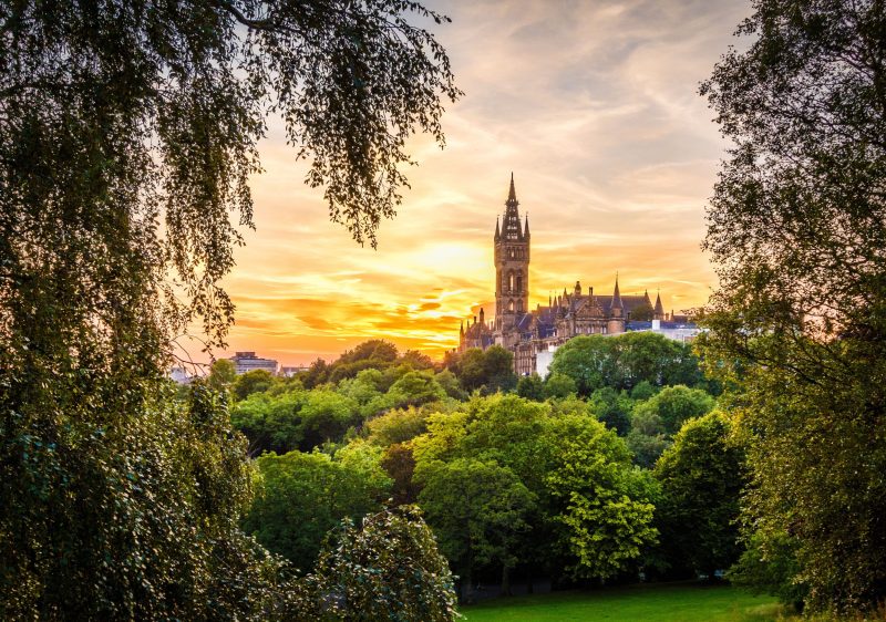 Sunset view of a tall, historic building with ornate architecture, surrounded by lush green trees. The foreground features leafy branches framing the scene, with a vibrant orange and yellow sky in the background.
