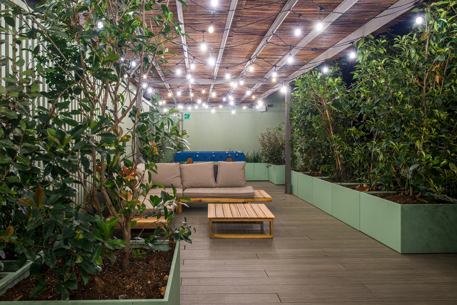 Outdoor patio with wooden flooring and potted plants along the sides. A canopy with string lights hangs overhead, creating a cozy atmosphere. A cushioned sofa and wooden coffee table are placed in the center. An exit sign is visible in the background.