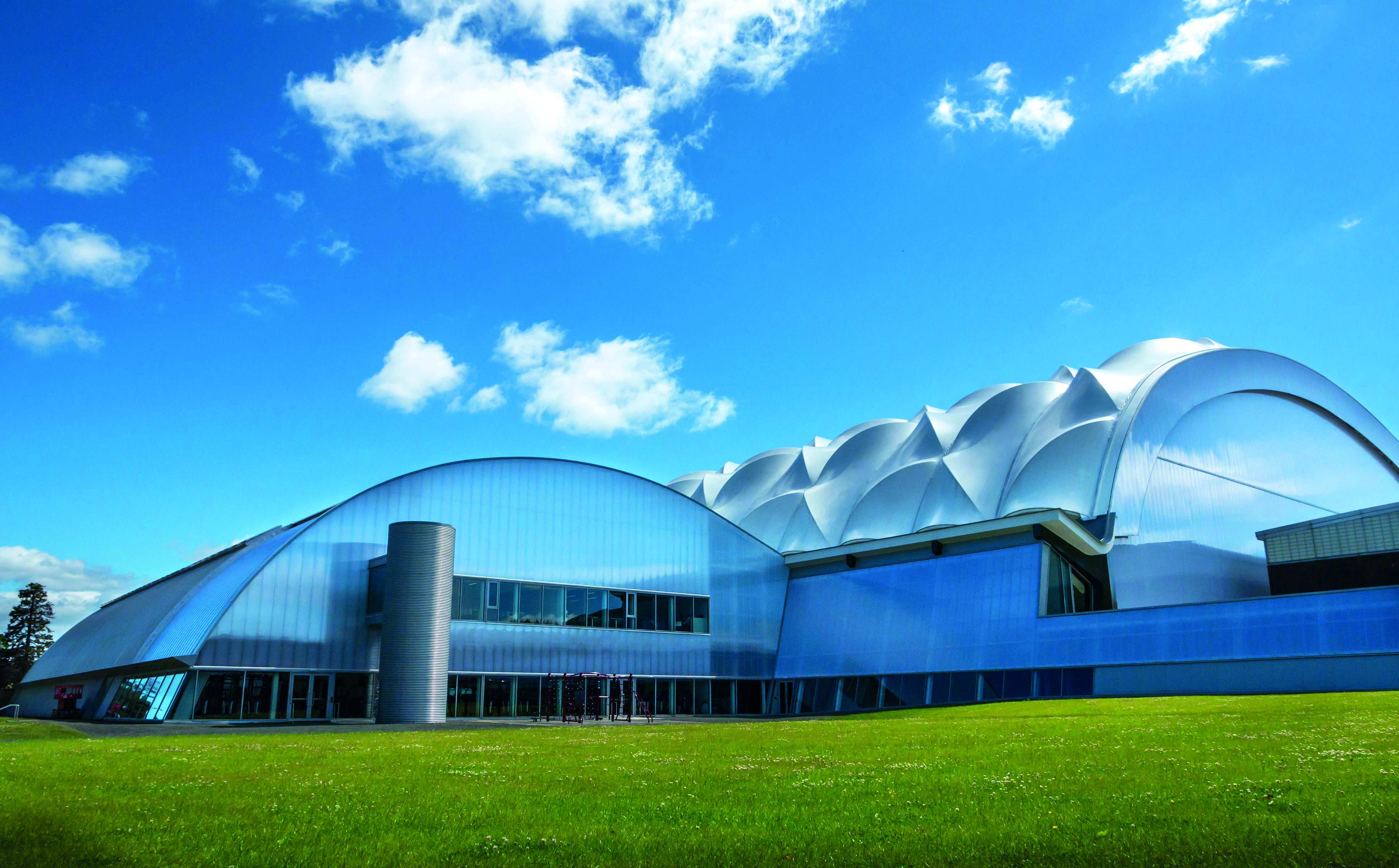 A modern building with a unique curved and domed architecture, featuring metallic surfaces and large glass windows. It is set against a bright blue sky with scattered clouds and surrounded by a green lawn.