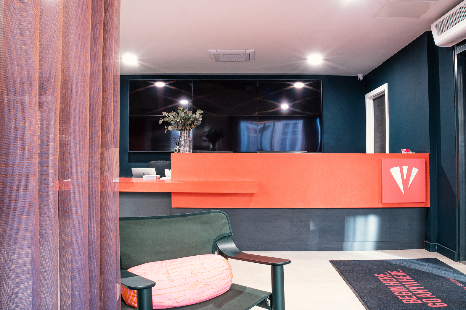 A modern office reception area with a bright red desk and a black wall behind it. A black chair with a pink cushion sits in the foreground, next to a semi-transparent curtain. The flooring is dark, and a small sign with text is on the floor by the entrance.