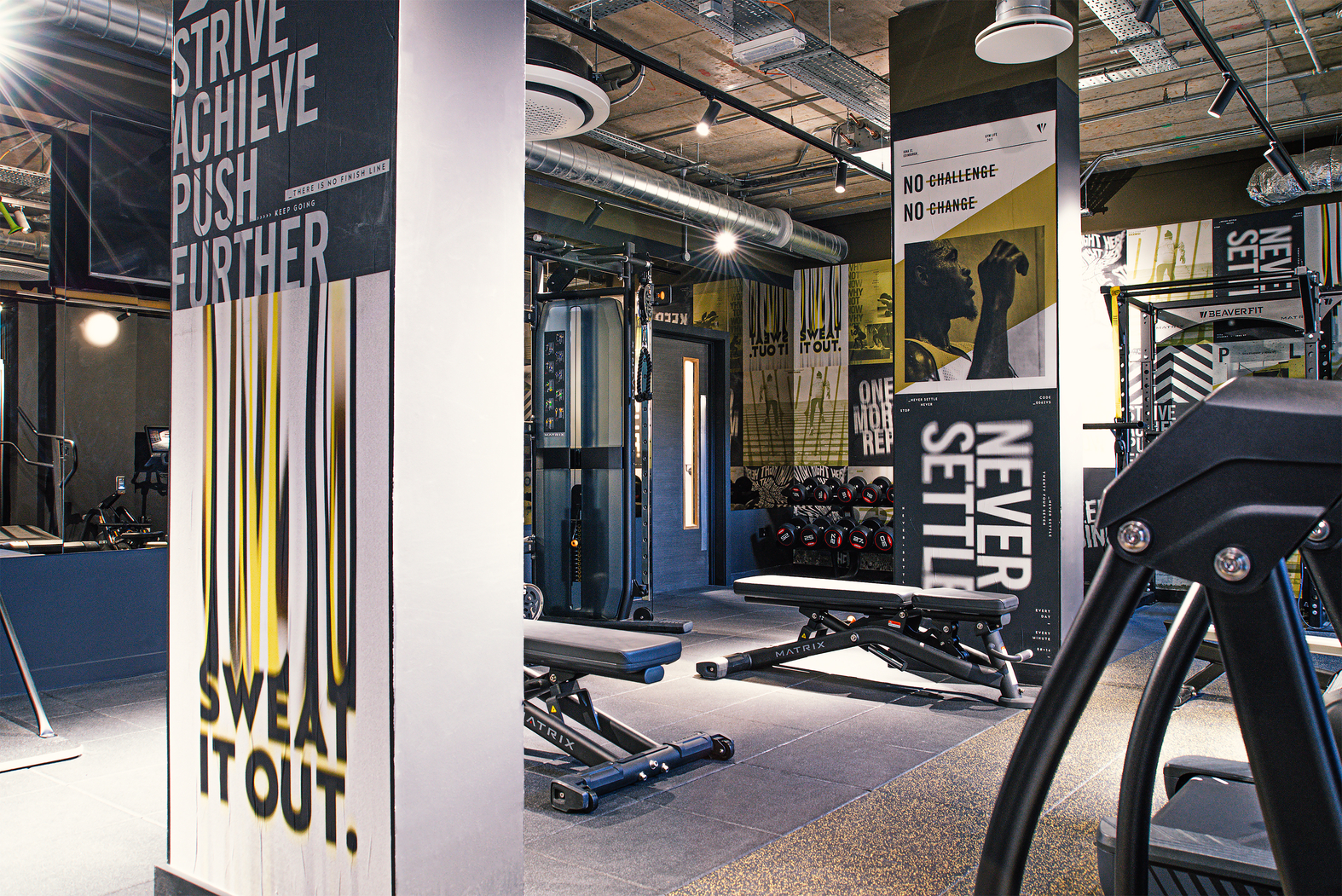 A modern gym with motivational posters on the walls, featuring messages like "Strive Achieve Push Further" and "Never Settle." The space includes benches, weight racks, and exercise machines. The lighting is bright, creating an energetic atmosphere.