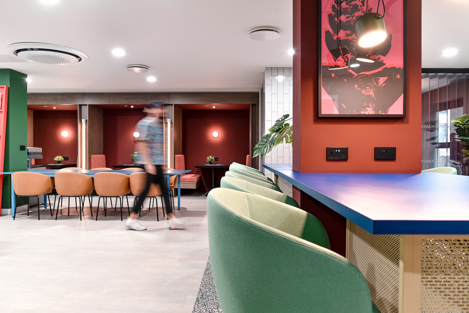 Modern interior with vibrant colors featuring a blue counter with green chairs, a blurred figure walking, orange chairs around tables in the background, red walls, artwork, and potted plants. Ceiling lights illuminate the space.