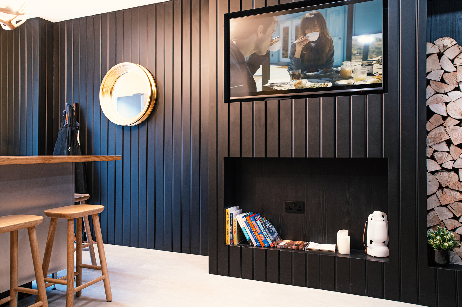 Bar space with wooden stools, dark vertical-paneled walls, and a round mirror. A TV displays a scene of two people eating. A niche holds books and small decorative items. Wood logs are neatly stacked nearby. Bright lighting enhances the modern aesthetic.