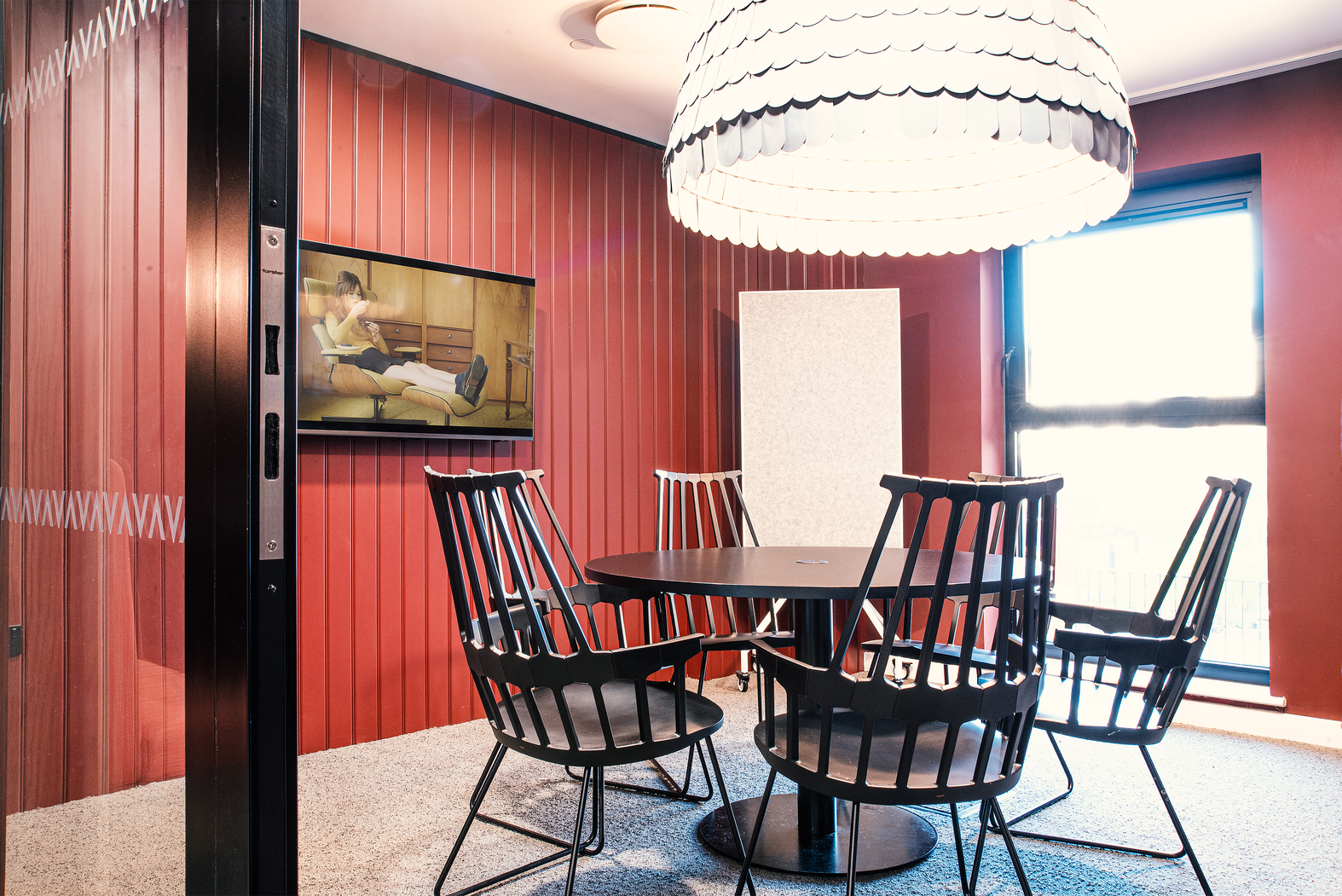 A modern meeting room with red walls, featuring a round black table surrounded by black chairs. A large, white, dome-shaped light fixture hangs above. A wall-mounted TV is on the side, and there's a window with blinds partially closed.