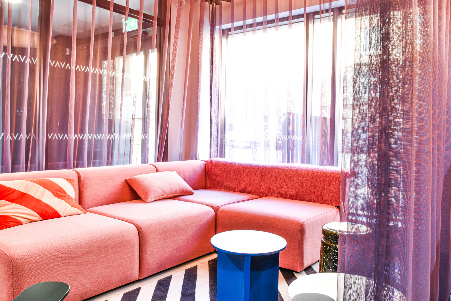 A modern living room with a pink sectional sofa and cushions. Sheer pink curtains hang by large windows, allowing natural light. A blue cylindrical table and black-and-white striped rug complete the vibrant decor.