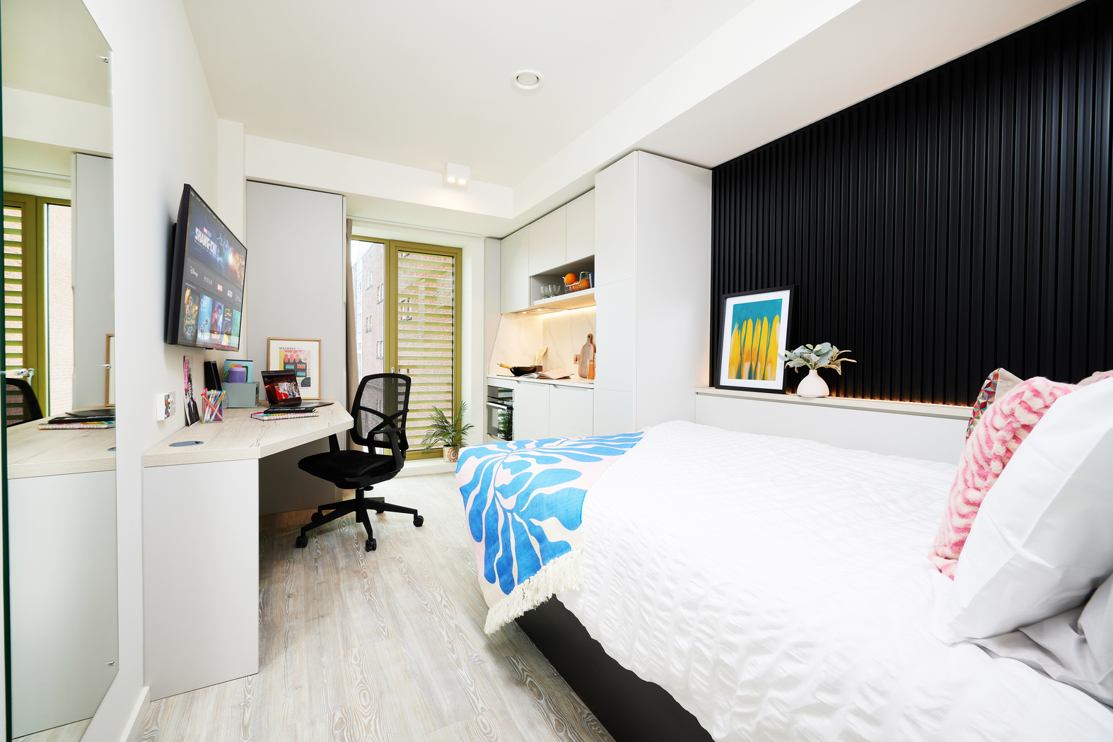 Modern, bright studio apartment on Bruce Street with a cozy bed, TV, desk, and office chair. The vibrant blue and white throw accents the bed perfectly. Kitchenette with wooden cabinets sits in the corner, while a window with blinds lets in natural light. Minimalist decor completes this charming space.