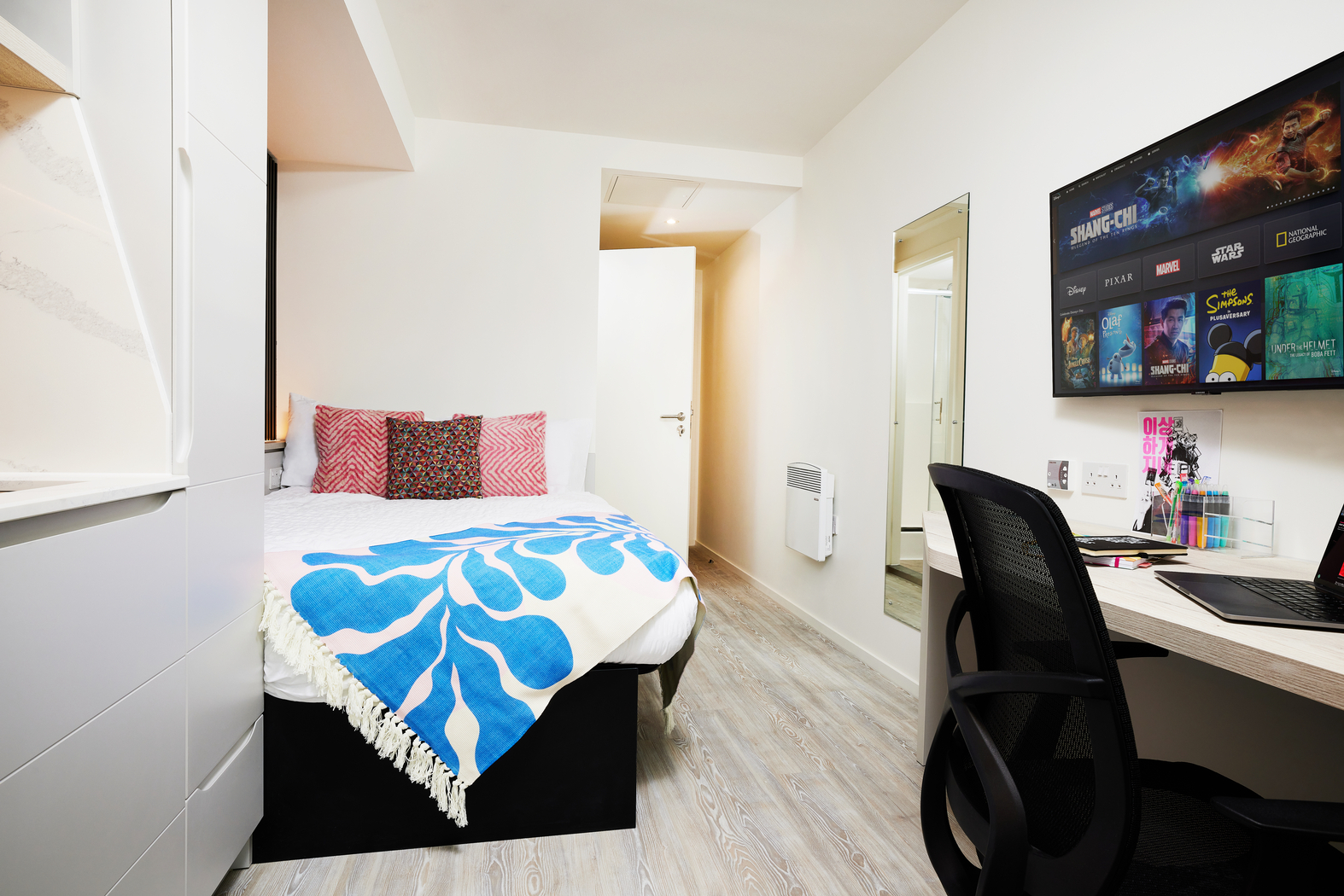 A tidy bedroom on Bruce Street showcases a bed with colorful pillows and a blue patterned throw. A desk with a chair sits opposite, featuring a laptop. A wall-mounted TV displays various movie posters. The room boasts minimalist and modern decor with light wood flooring.