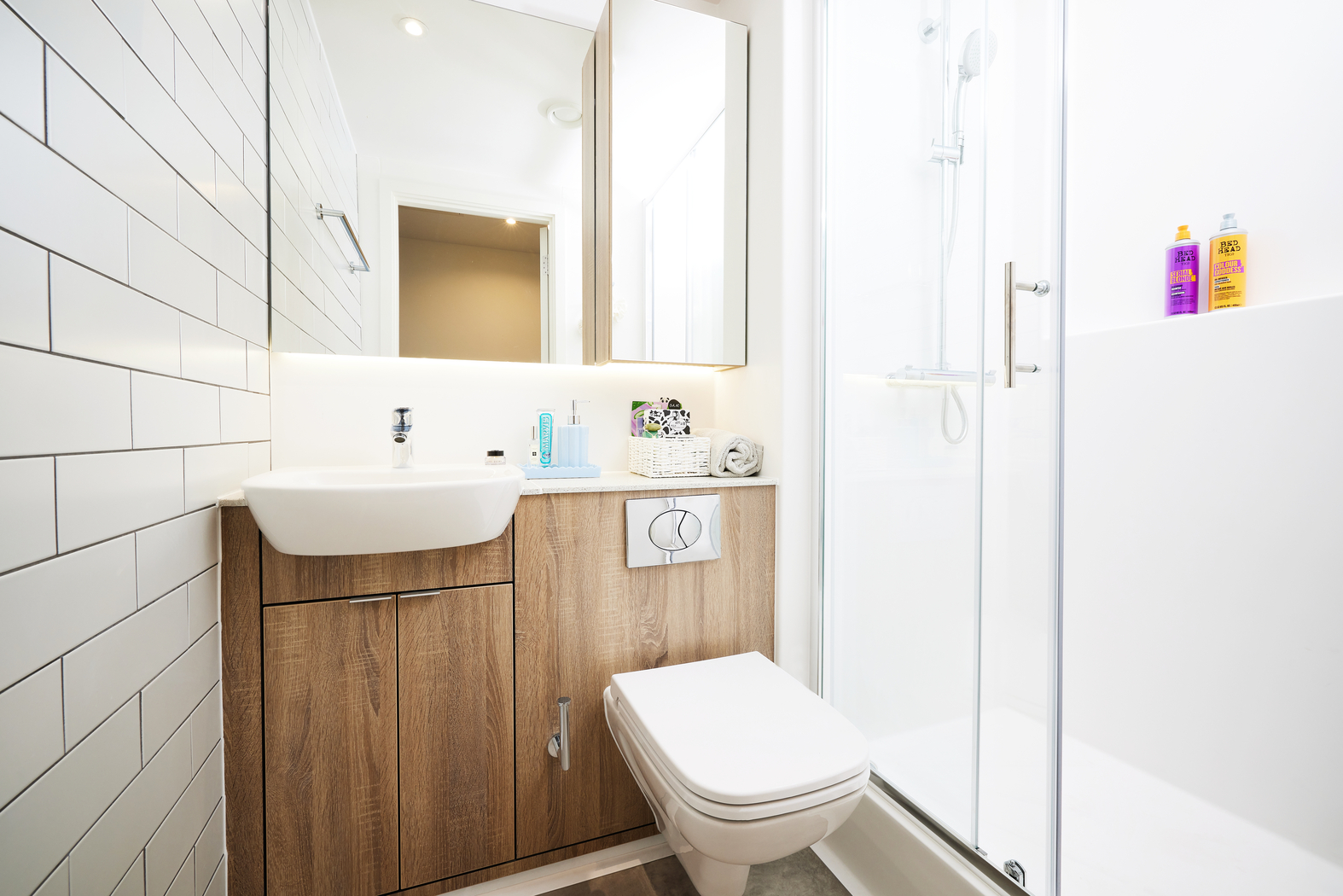 A modern bathroom on Bruce Street features a white wall-mounted sink and toilet, a wooden vanity, a large mirror, and a glass-enclosed shower. The white tiled walls and bright lighting enhance the space's clean, contemporary feel.