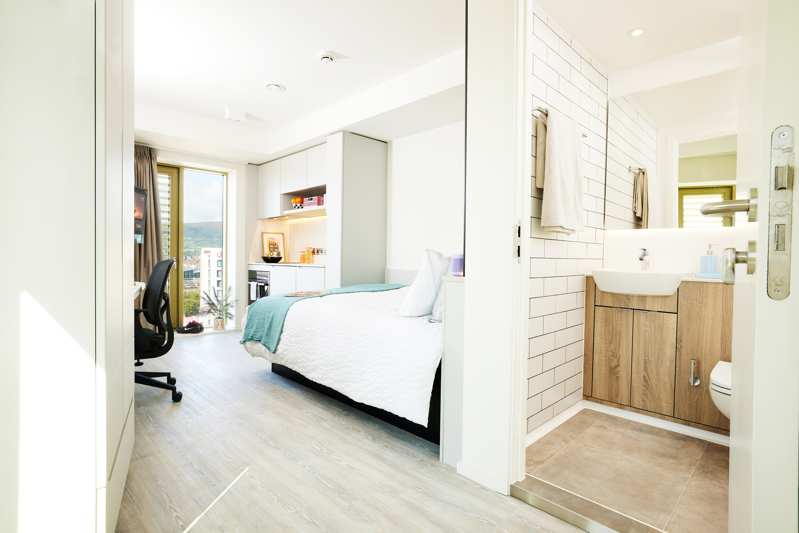A bright, modern studio apartment on Bruce Street features a cozy bed, desk with chair, and large window. The bathroom boasts white tiled walls and a wooden vanity with a sink. Light wood flooring throughout enhances the airy feel.
