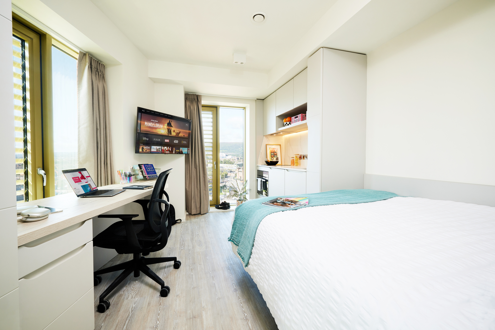A tidy, modern bedroom on Bruce Street features a bed, desk, and office chair. A laptop and snacks sit on the desk, while a mounted TV showcases streaming services. A large window provides views of the cityscape. Soft lighting enhances the cozy atmosphere.