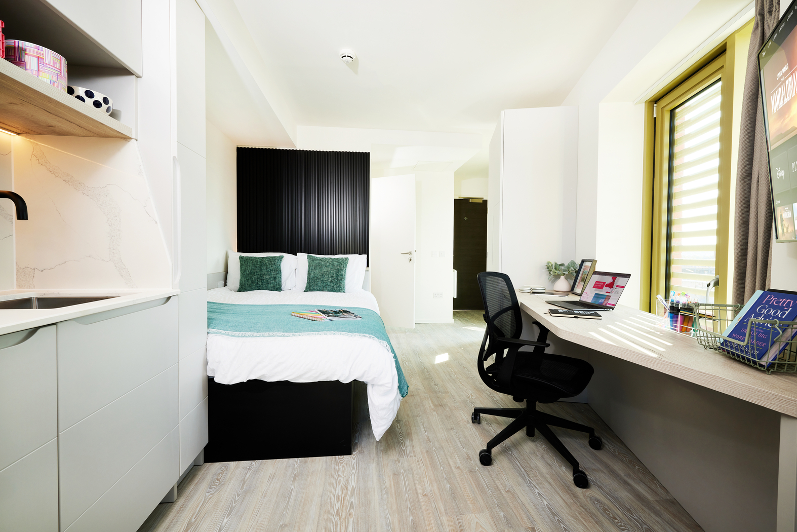 A modern studio apartment on Bruce Street features a cozy bed and teal accents. A spacious desk with a laptop and books sits beside a large window with blinds. The kitchenette, with white cabinets, is on the left, and the room boasts light wood flooring.