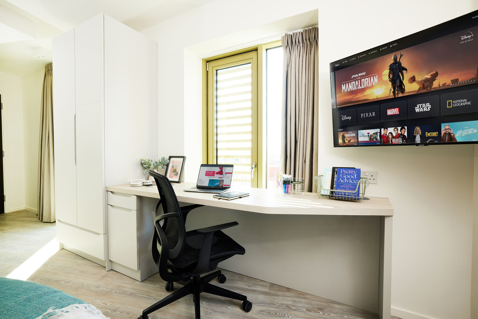 Located on Bruce Street, this modern home office features a curved desk, black office chair, and wall-mounted TV showcasing streaming services. A laptop and stationery adorn the desk, while a white wardrobe and gray-curtained window complete the bright, minimalistic decor.